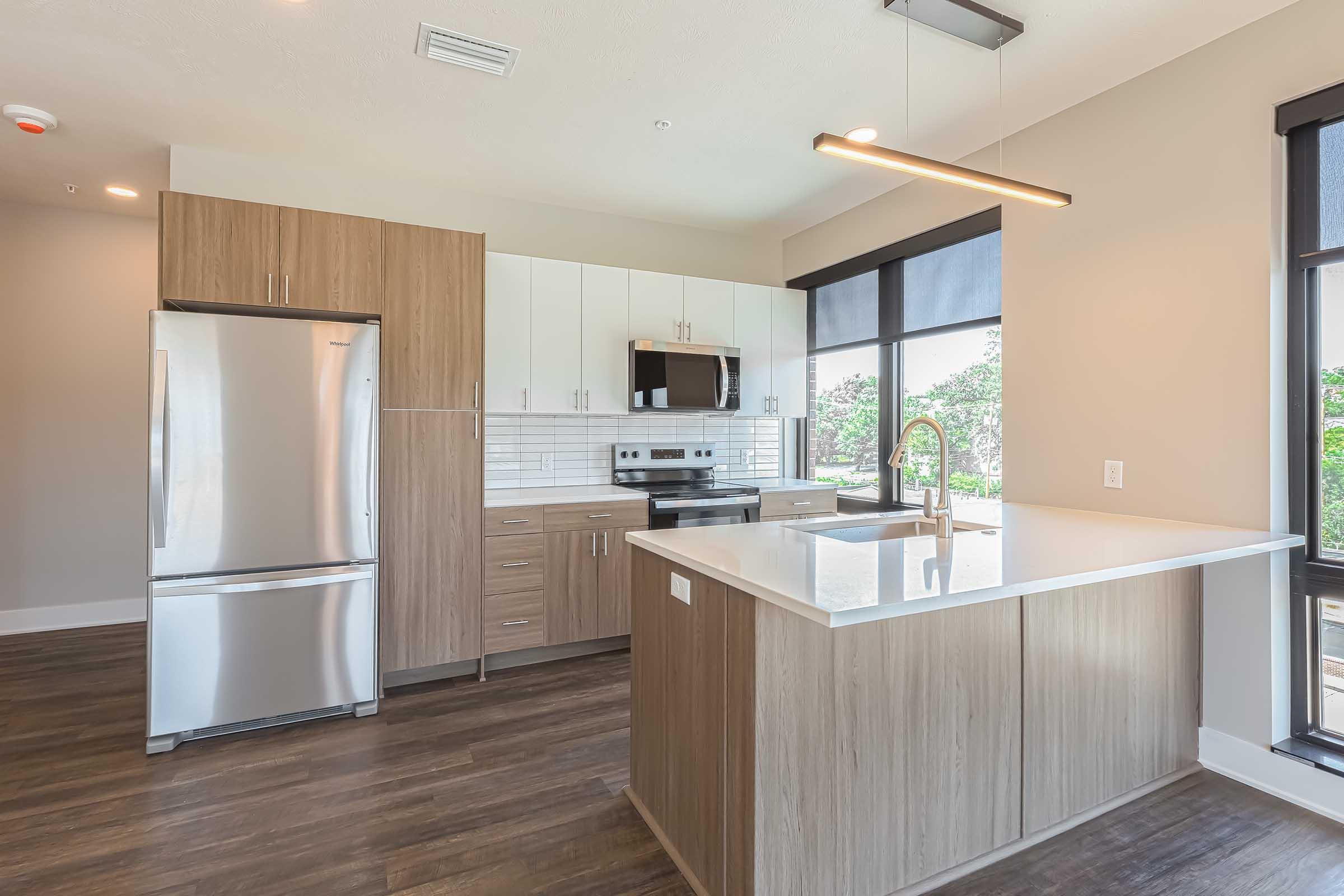 Modern kitchen with a stainless steel fridge, microwave, and stove. Features light wood cabinetry, a white countertop, and a sink. Large windows provide natural light, and the floor is dark wood. The design is sleek and contemporary, making the space feel open and inviting.