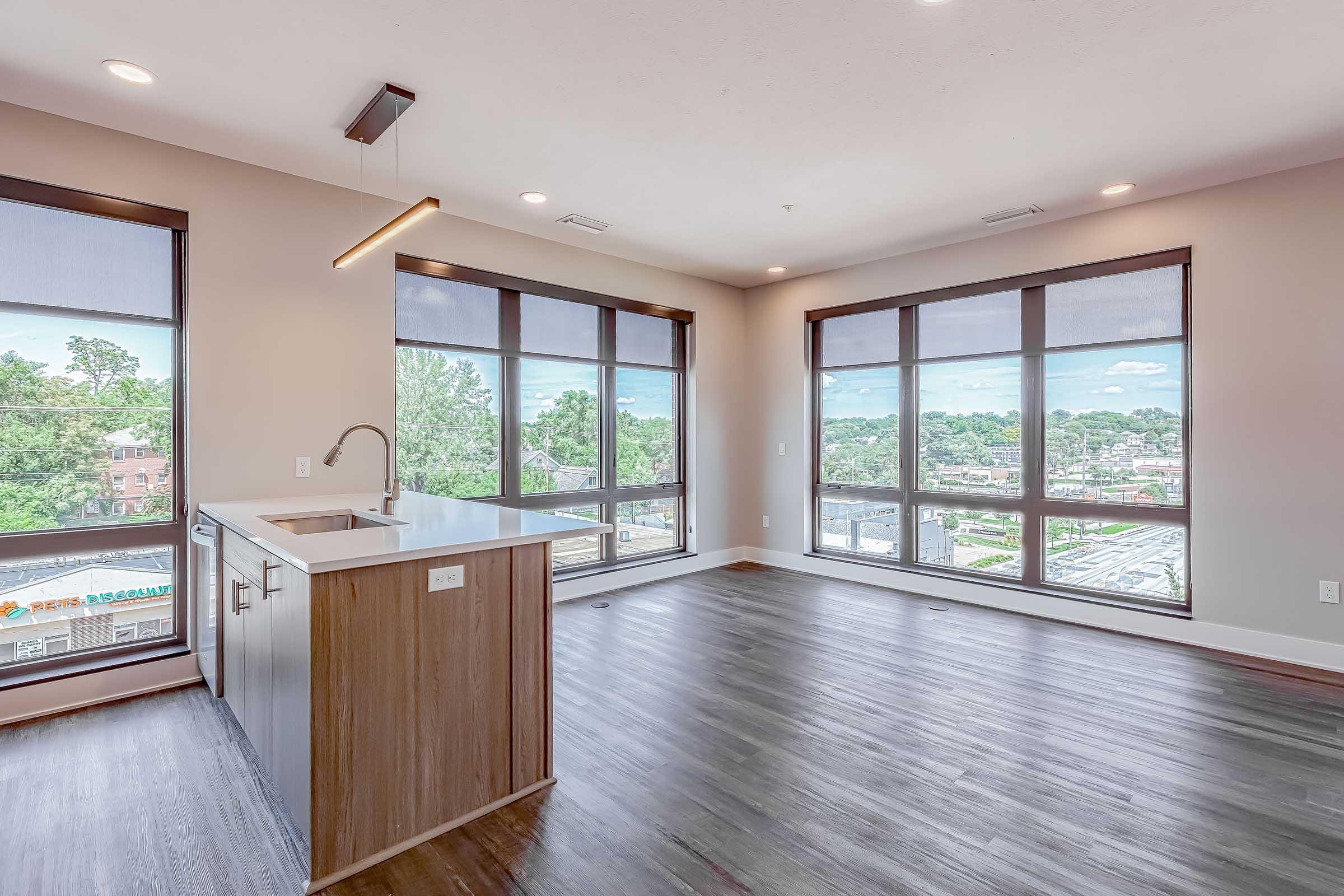 A modern, empty apartment with large windows allowing natural light. The kitchen area features a small island and sleek cabinetry. The flooring is dark wood, and the walls are painted a light color. Outside the windows, there is a view of greenery and city buildings.