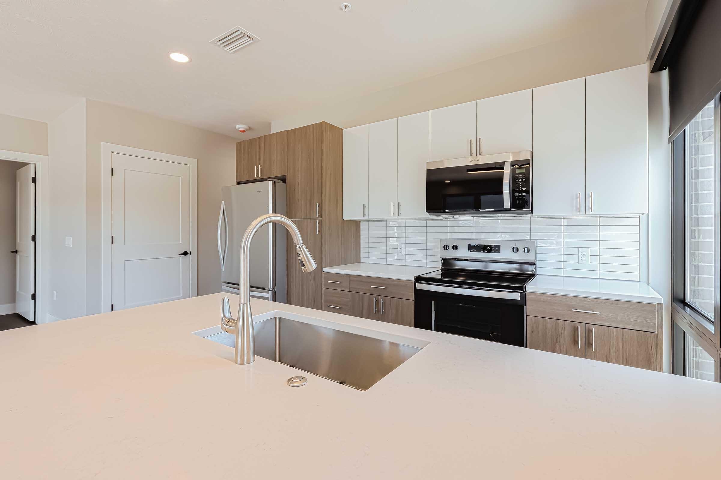 Modern kitchen featuring a large island with a stainless steel sink, sleek white cabinets, and wooden accents. The appliances include a microwave, oven, and refrigerator, all in stainless steel. Bright natural light comes through a window, highlighting the clean lines and contemporary design.