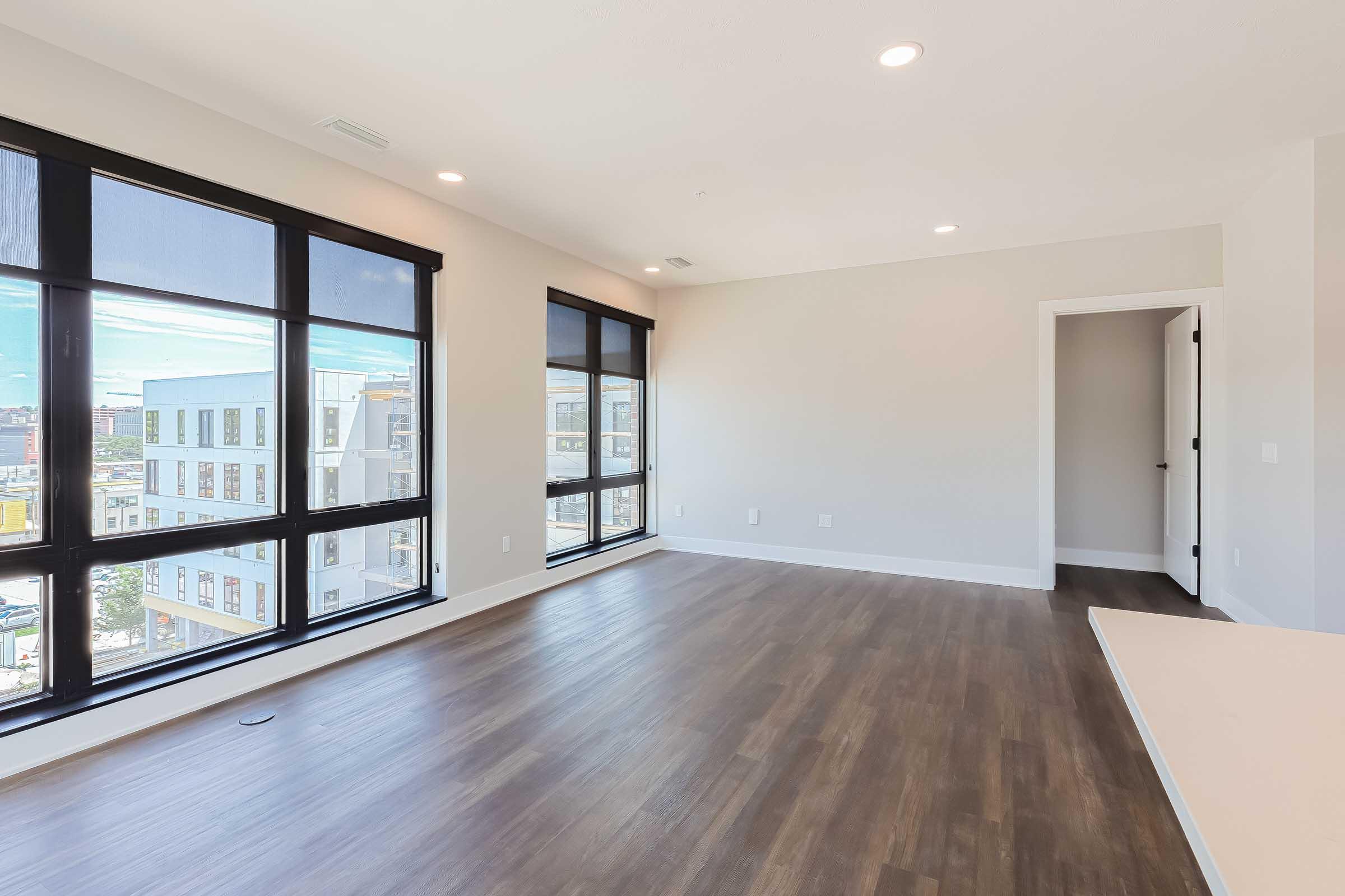 Spacious, modern apartment interior featuring large windows with black roller shades, light-colored walls, and hardwood flooring. An open floor plan includes a view of the city skyline and a doorway leading to another room. Natural light floods the space, creating a bright and inviting atmosphere.