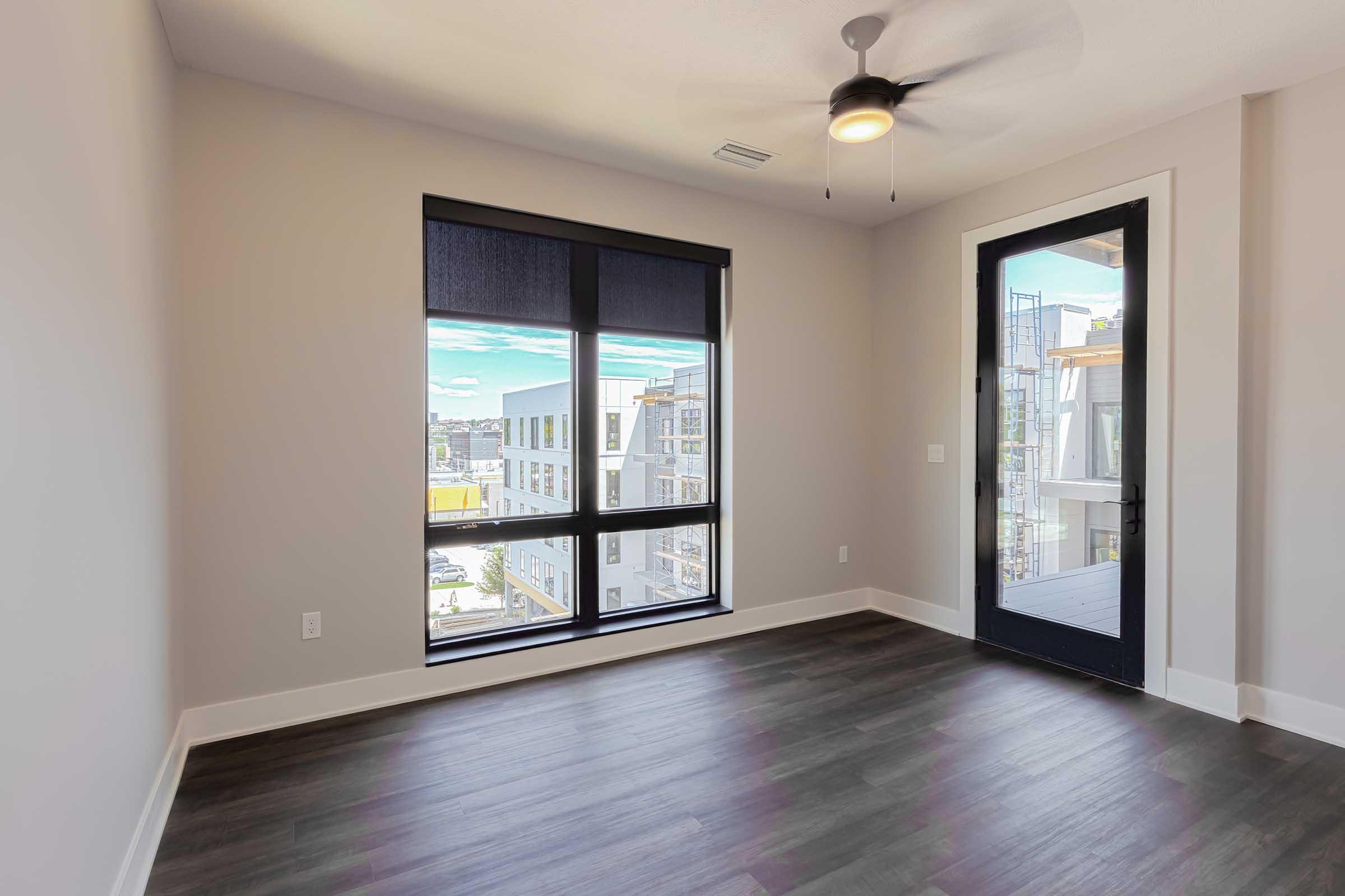 Spacious room with a ceiling fan, featuring large windows with blinds that offer a view of an urban landscape. The flooring is dark and modern, and there's a sliding door leading to a balcony area. The walls are light-colored, creating a bright and airy atmosphere.