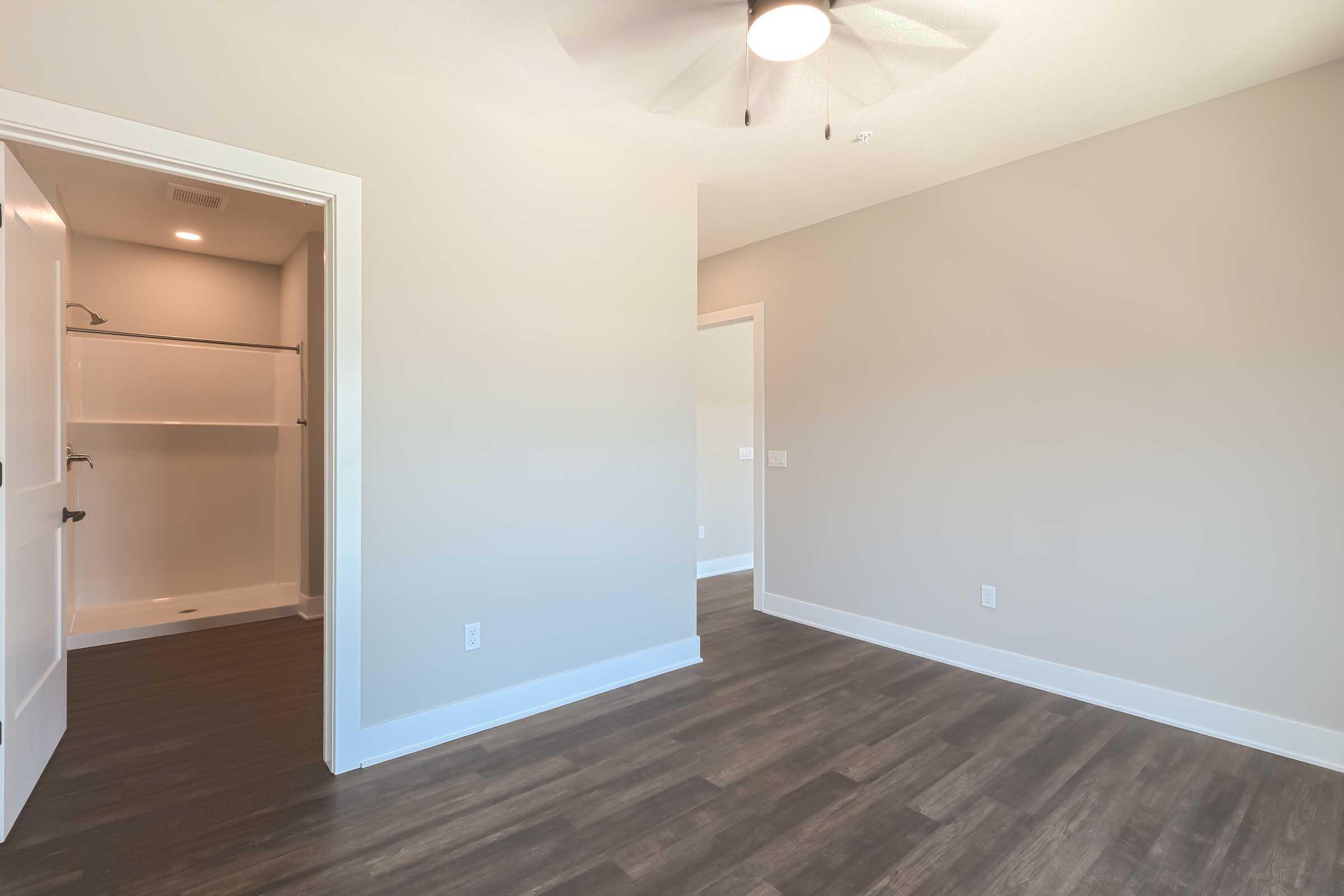 A spacious room with light gray walls, a ceiling fan, and dark laminate flooring. There is an opening leading to a closet with a shelf and a door to a bathroom area, which has a modern design. Natural light fills the space, creating a bright and inviting atmosphere.