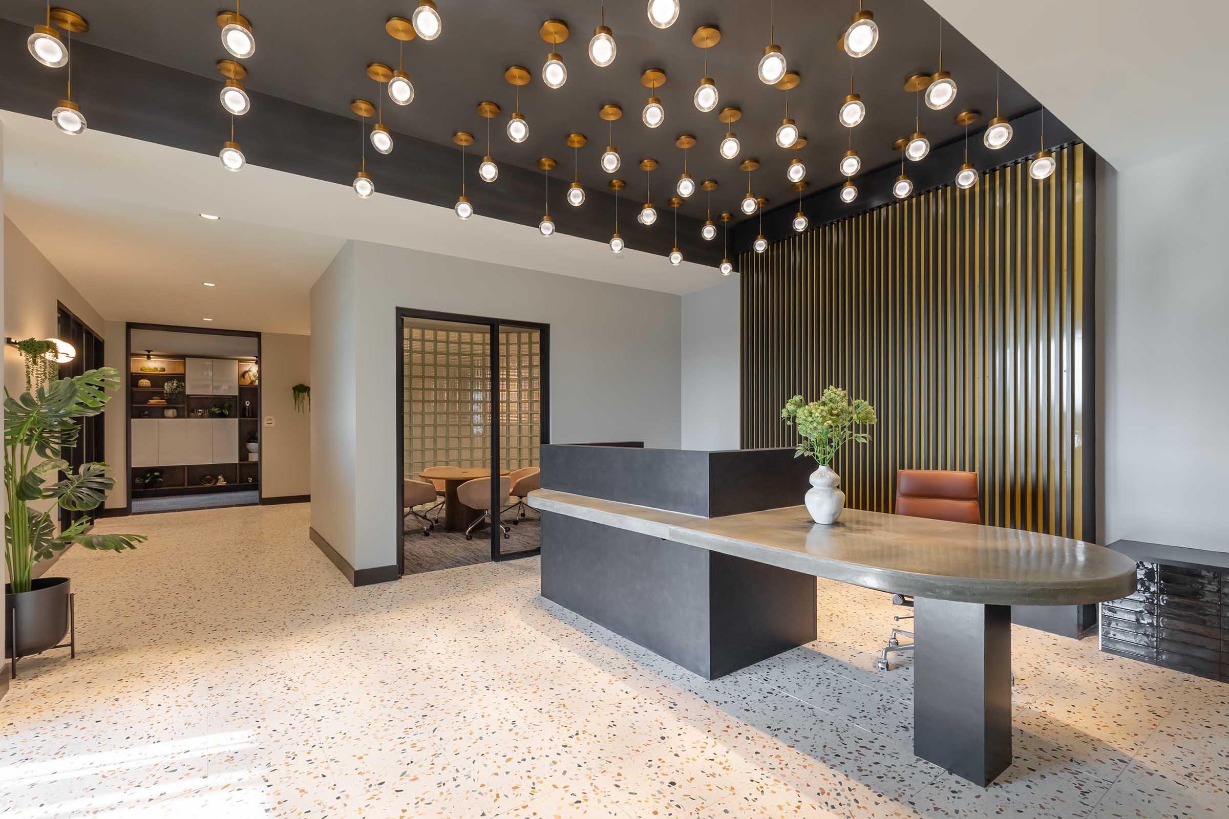 Modern office lobby featuring a sleek reception desk with a curved surface, decorative hanging light fixtures, and stylish wall paneling. The space includes potted plants and a seating area, all in a contemporary design with a mix of materials and textures.