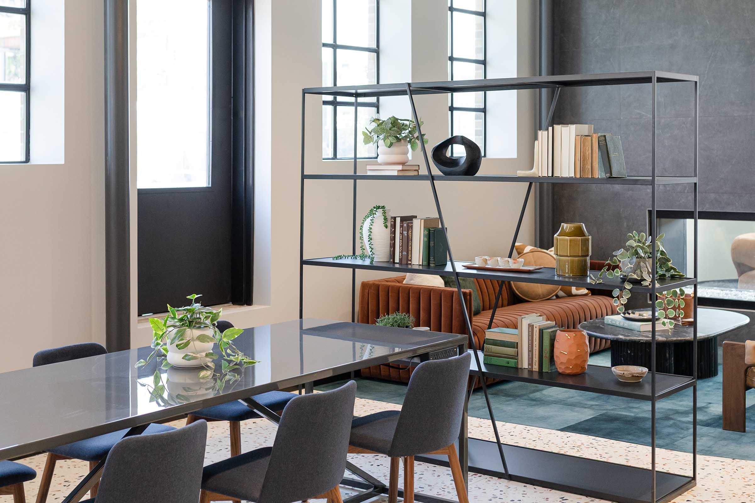 A modern dining space featuring a sleek black metal shelving unit filled with books and decorative items. A long dining table with upholstered chairs sits in front, while a cozy seating area with a brown couch and various plants adds warmth to the room. Large windows allow natural light to fill the space.