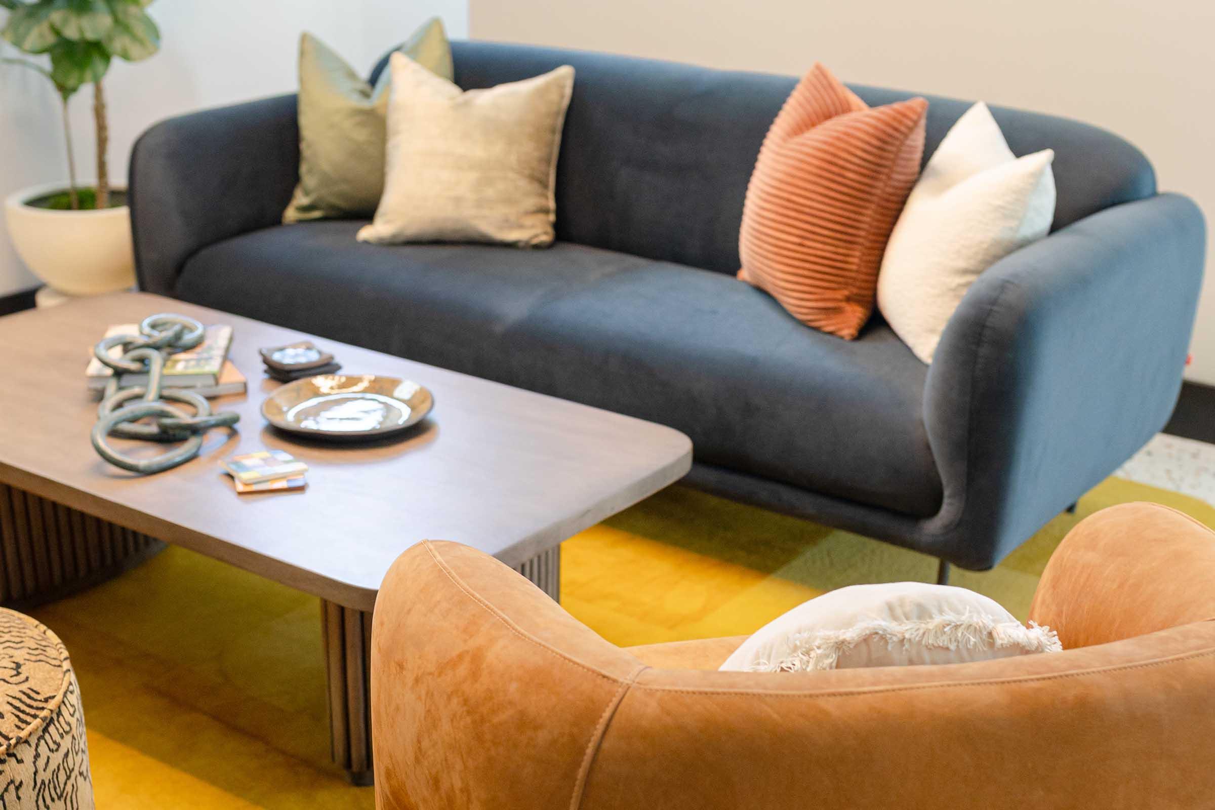 A cozy living room setup featuring a navy blue sofa adorned with various decorative pillows in neutral tones, a wooden coffee table with magazines and an ashtray, and a stylish armchair in a warm beige color. The space is enhanced by a potted plant and a brightly colored area rug.