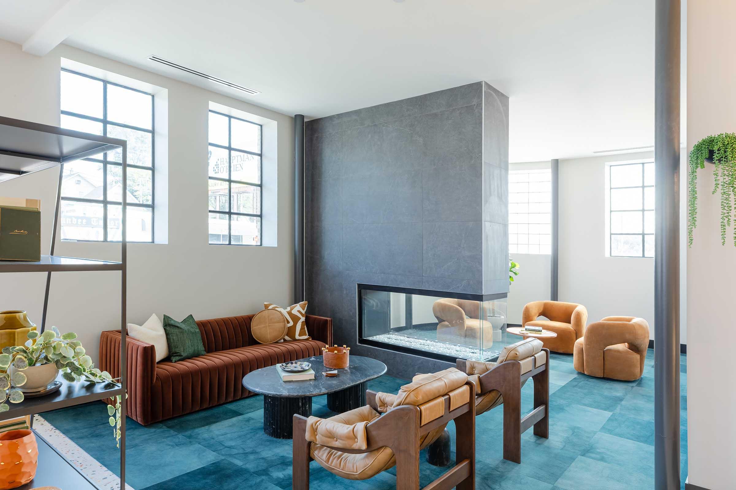 A modern living space featuring a sleek fireplace, a cozy brown sofa with vertical stripes, and stylish armchairs. The room has large windows allowing ample natural light, a circular dark coffee table, and a blue-green rug, creating a warm and inviting atmosphere.