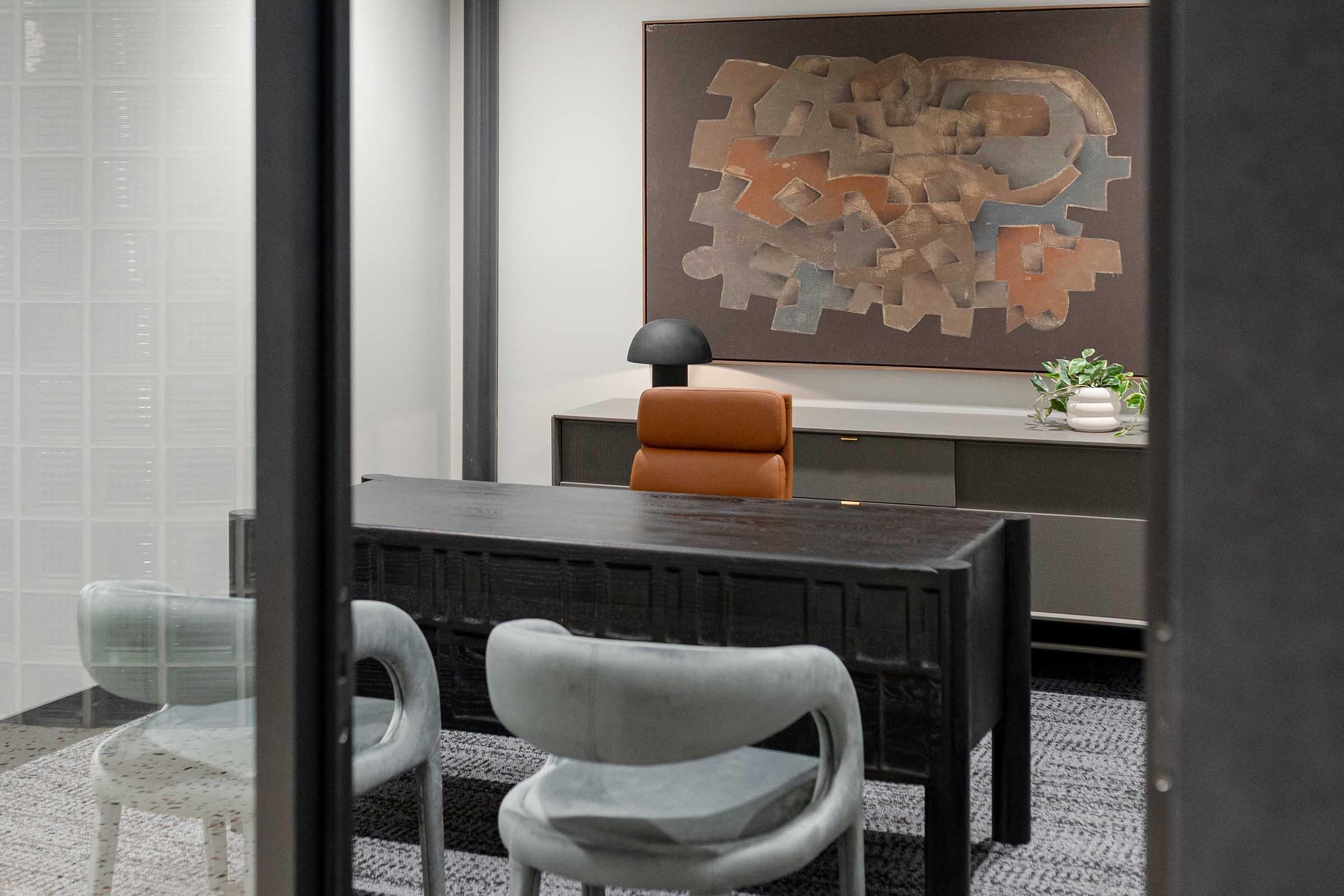 A modern office space featuring a wooden desk with an orange chair, two light-colored chairs, and a minimalistic lamp. Behind the desk is a large abstract artwork on the wall. The room has glass walls, providing a glimpse into the well-lit interior decorated with neutral tones and a small potted plant.