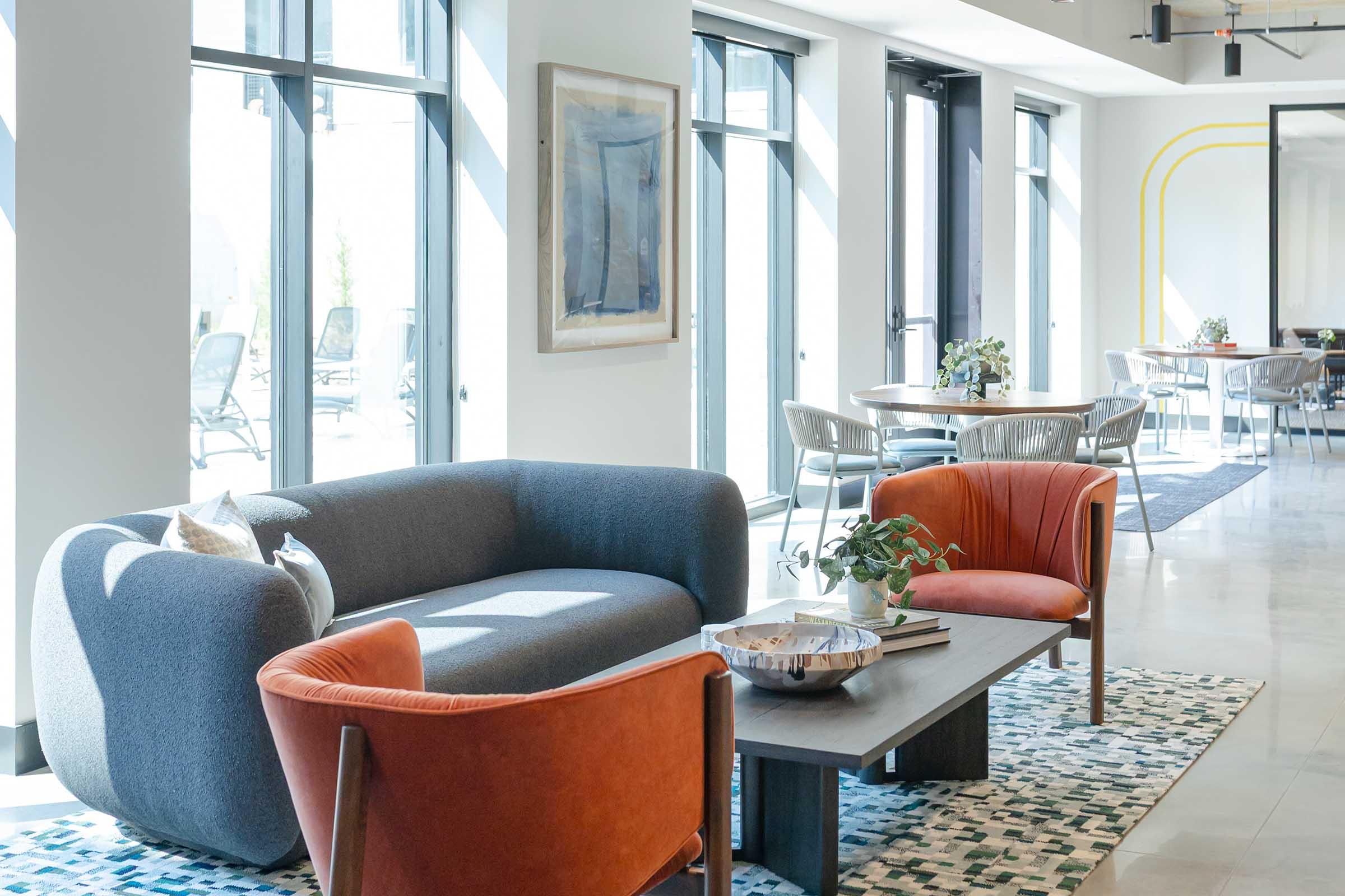 A modern lounge area featuring a gray curved sofa, two orange chairs, a wooden coffee table, and a decorative bowl. Large windows allow natural light to fill the space, which includes a dining area in the background with tables and chairs. A colorful rug adds texture to the floor.