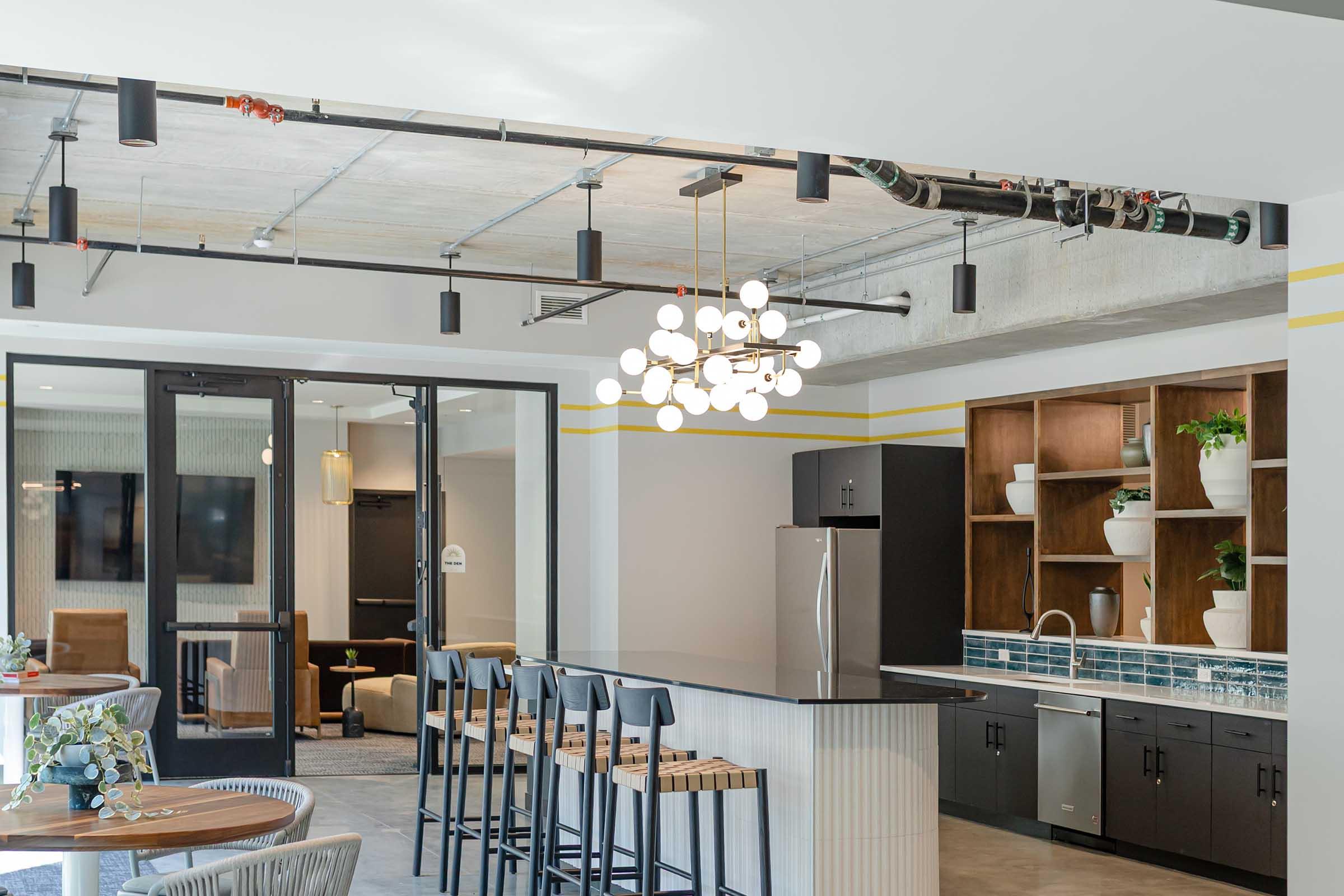 A modern kitchen area with a sleek black countertop and bar stools under pendant lighting. The background features a kitchenette with stainless steel appliances and wood shelving displaying plants. Large glass doors lead to a lounge area, with neutral-toned furniture visible in the distance.