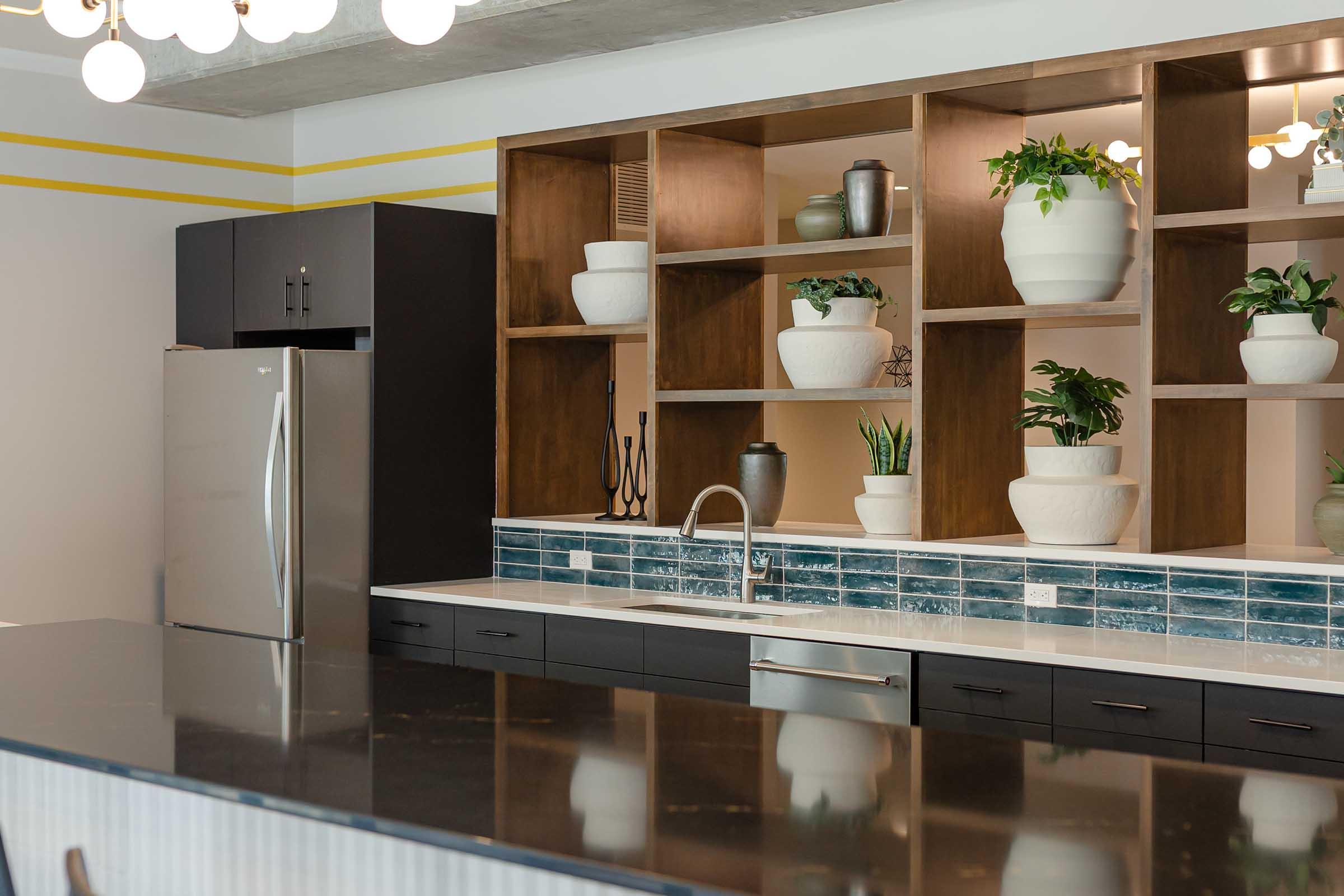 Modern kitchen featuring an open shelving unit with potted plants, decorative vases, and a minimalist design. A stainless steel refrigerator is on the left, while the island in the foreground has a dark countertop. The backsplash is accented with blue tiles, and there's a sleek faucet above the sink.