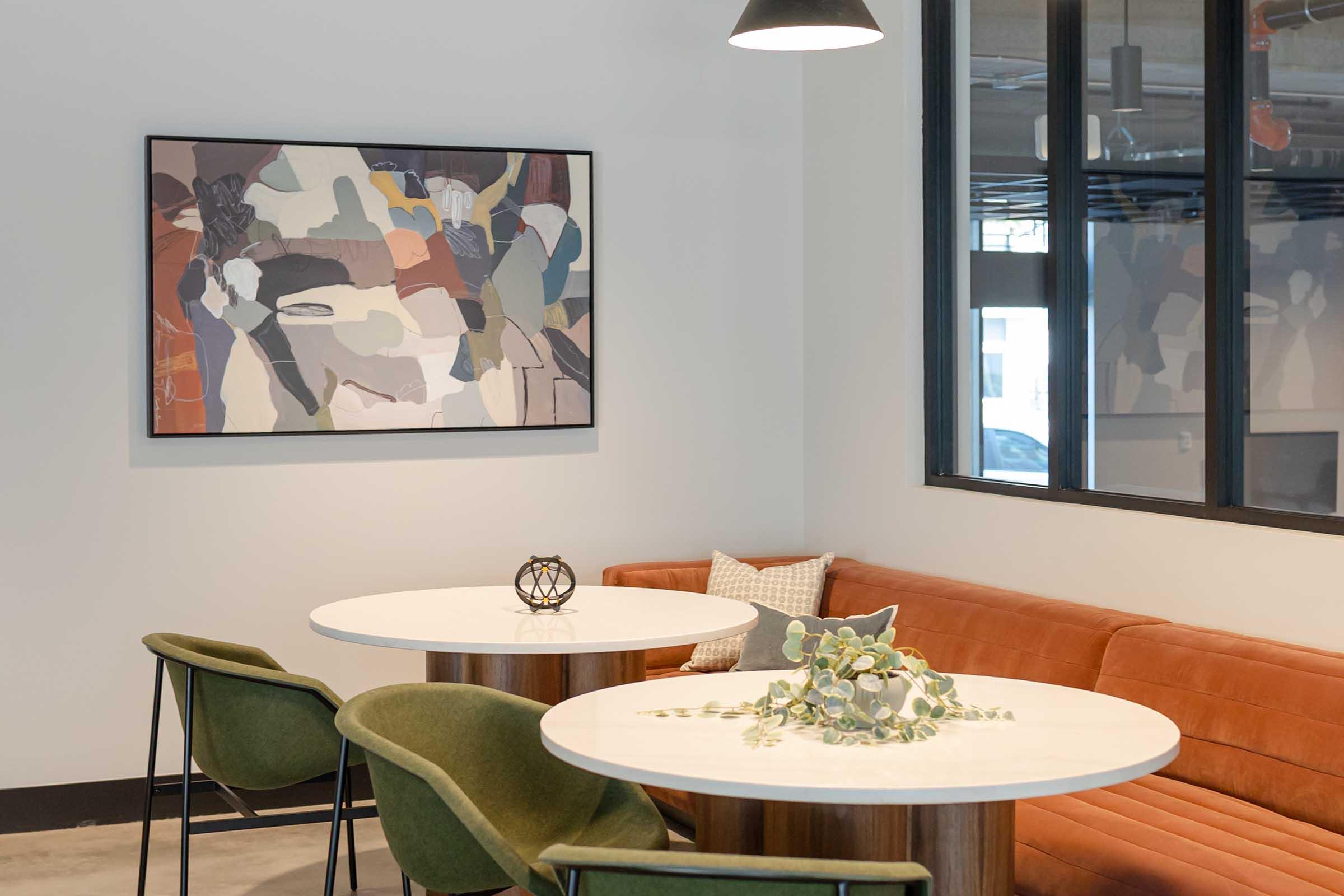 A modern lounge area featuring two round tables with light wooden bases, surrounded by green chairs. A warm, orange-brown couch is against the wall, adorned with a decorative pillow. Above the couch, there is a large abstract art piece with earthy tones. The space has large windows allowing natural light.