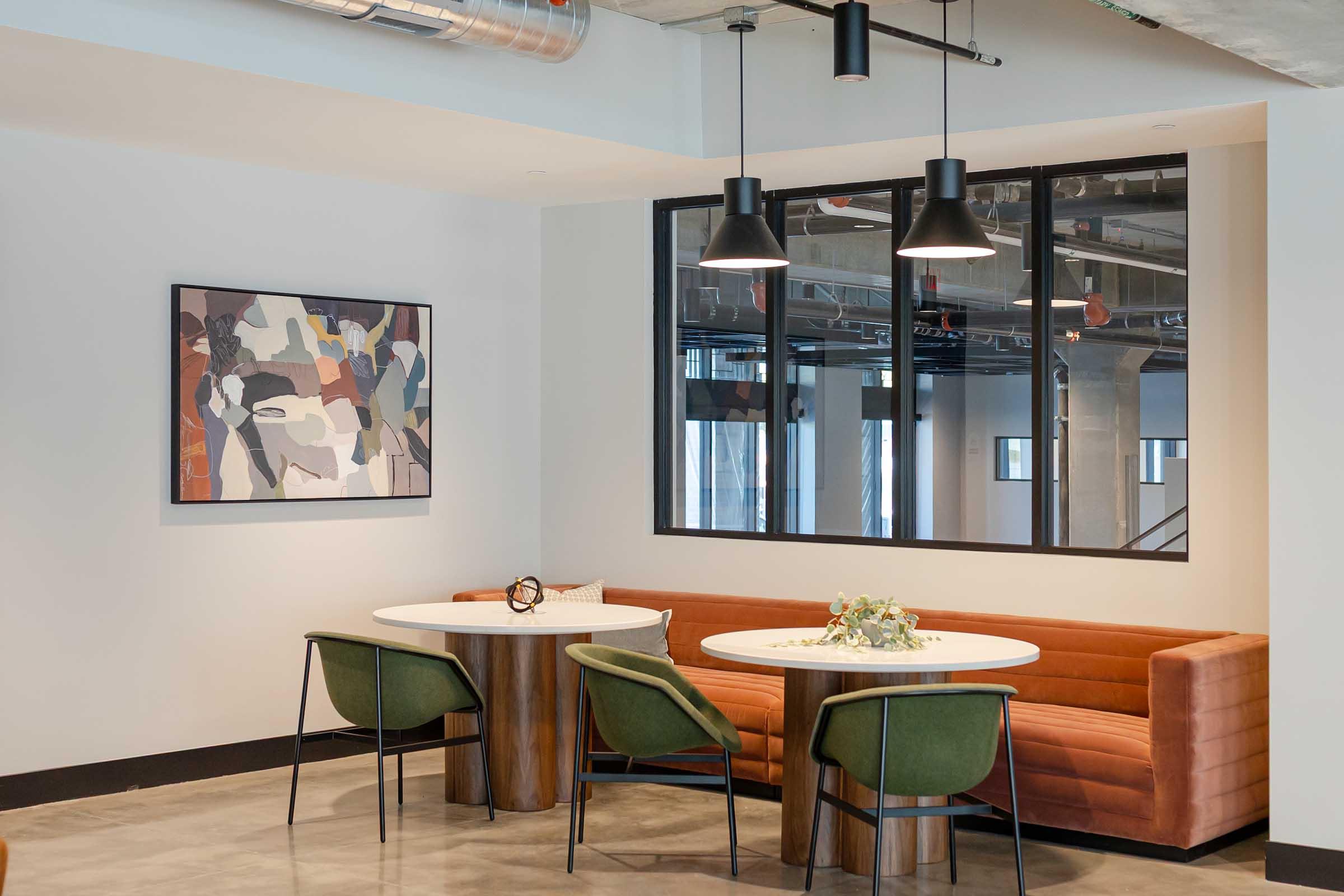 A modern interior space featuring a round table with a light wood finish, surrounded by green chairs. A stylish brown couch lines the wall, accompanied by a decorative vase of flowers. A large window allows natural light, and an abstract painting hangs on the wall, enhancing the contemporary aesthetic.