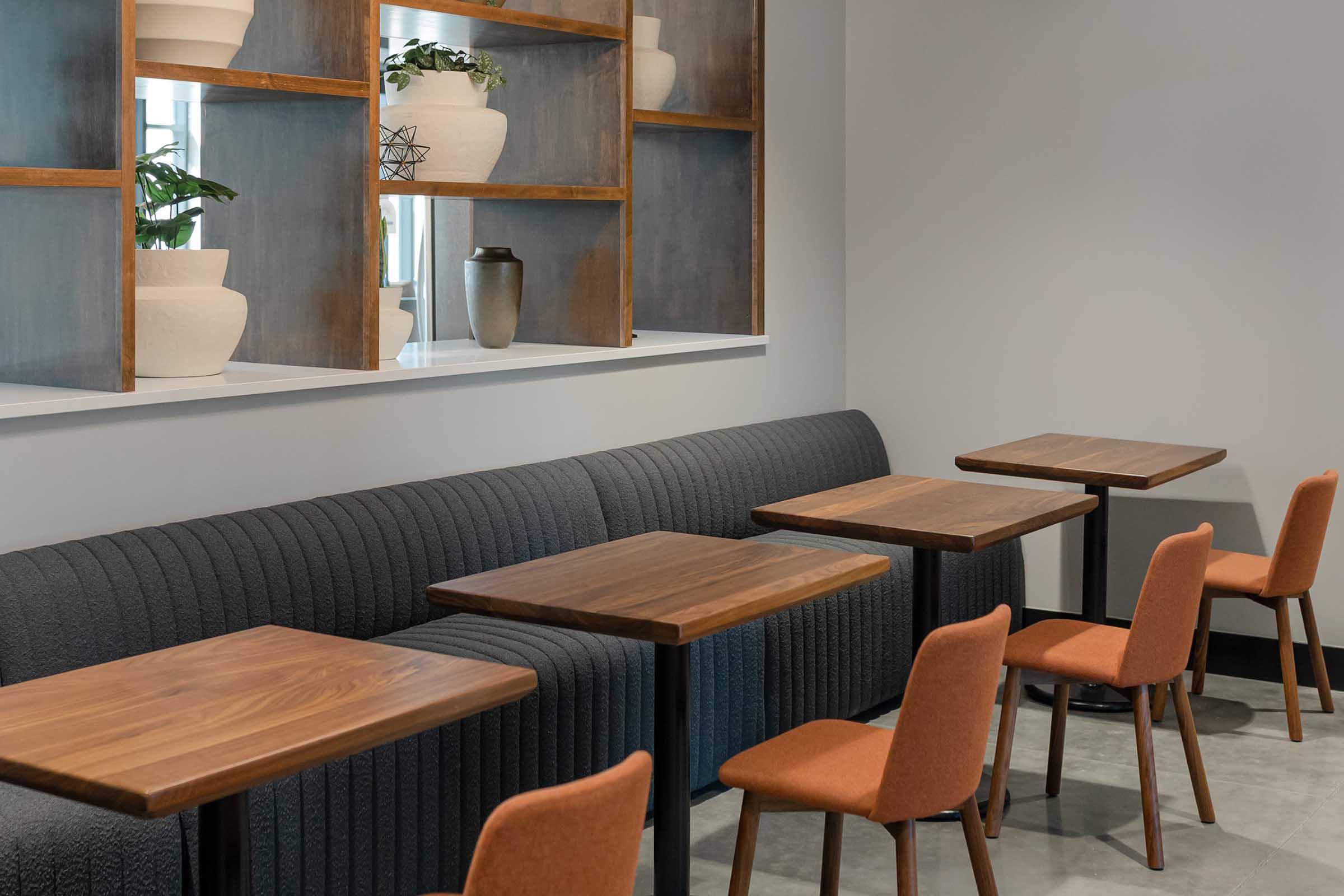 A modern café interior featuring a gray upholstered bench along the wall, several wooden tables, and orange chairs. Decorative shelves with potted plants and ceramic objects are positioned above the bench, creating a clean and inviting atmosphere.