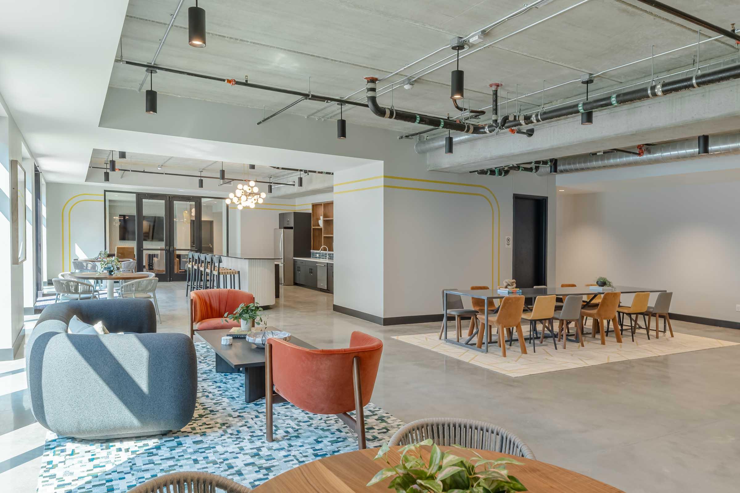 A modern open-concept workspace featuring a casual seating area with colorful chairs and sofas, a dining table surrounded by wooden chairs, and an adjacent kitchenette. The space has large windows allowing natural light and a contemporary design with industrial elements.