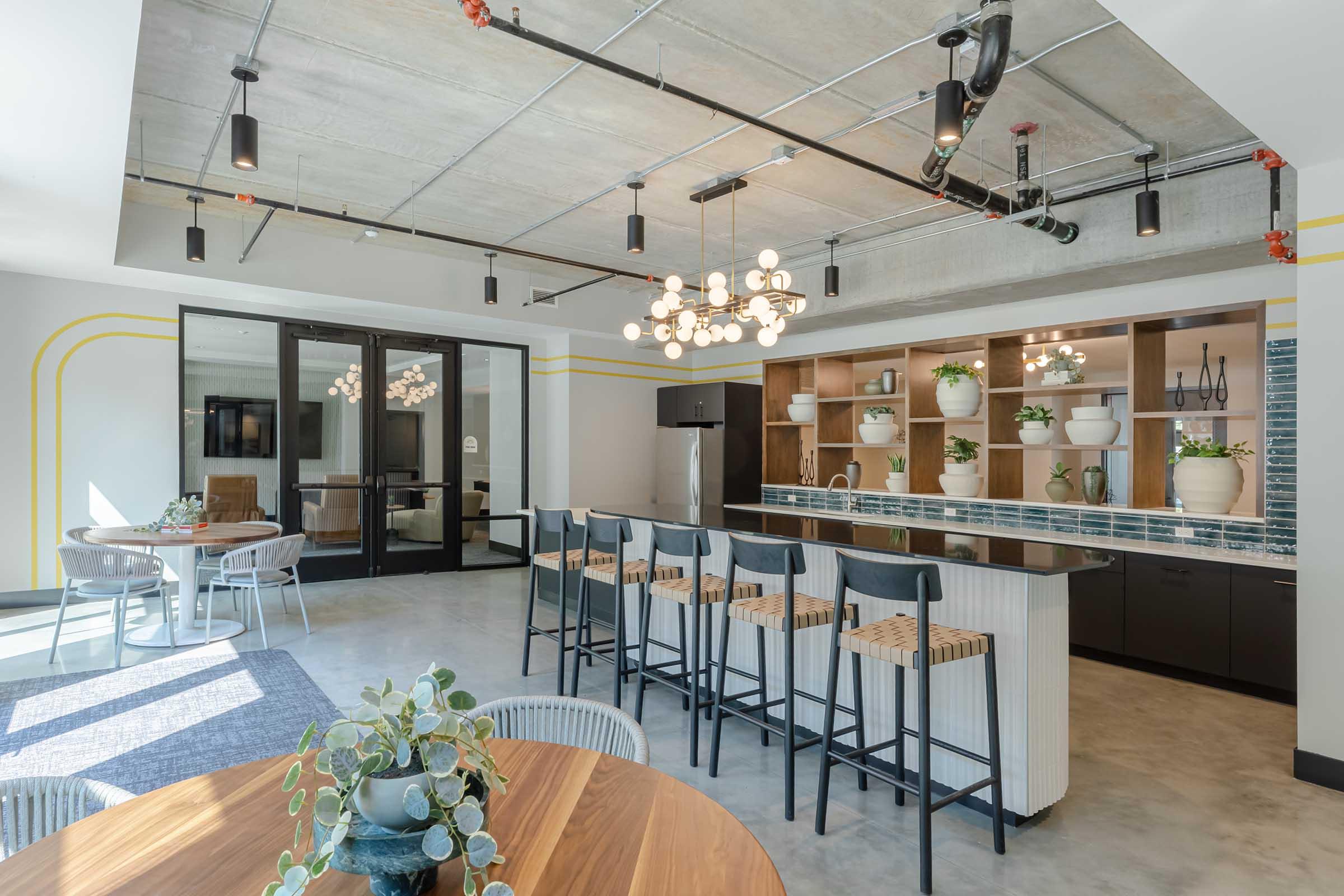 Modern communal kitchen with a long bar featuring stools, two round dining tables, and decorative plants. The space has a minimalist design with concrete floors, industrial lighting fixtures, and a large glass door leading to another room. A stylish backsplash and shelves with potted plants add a contemporary touch.