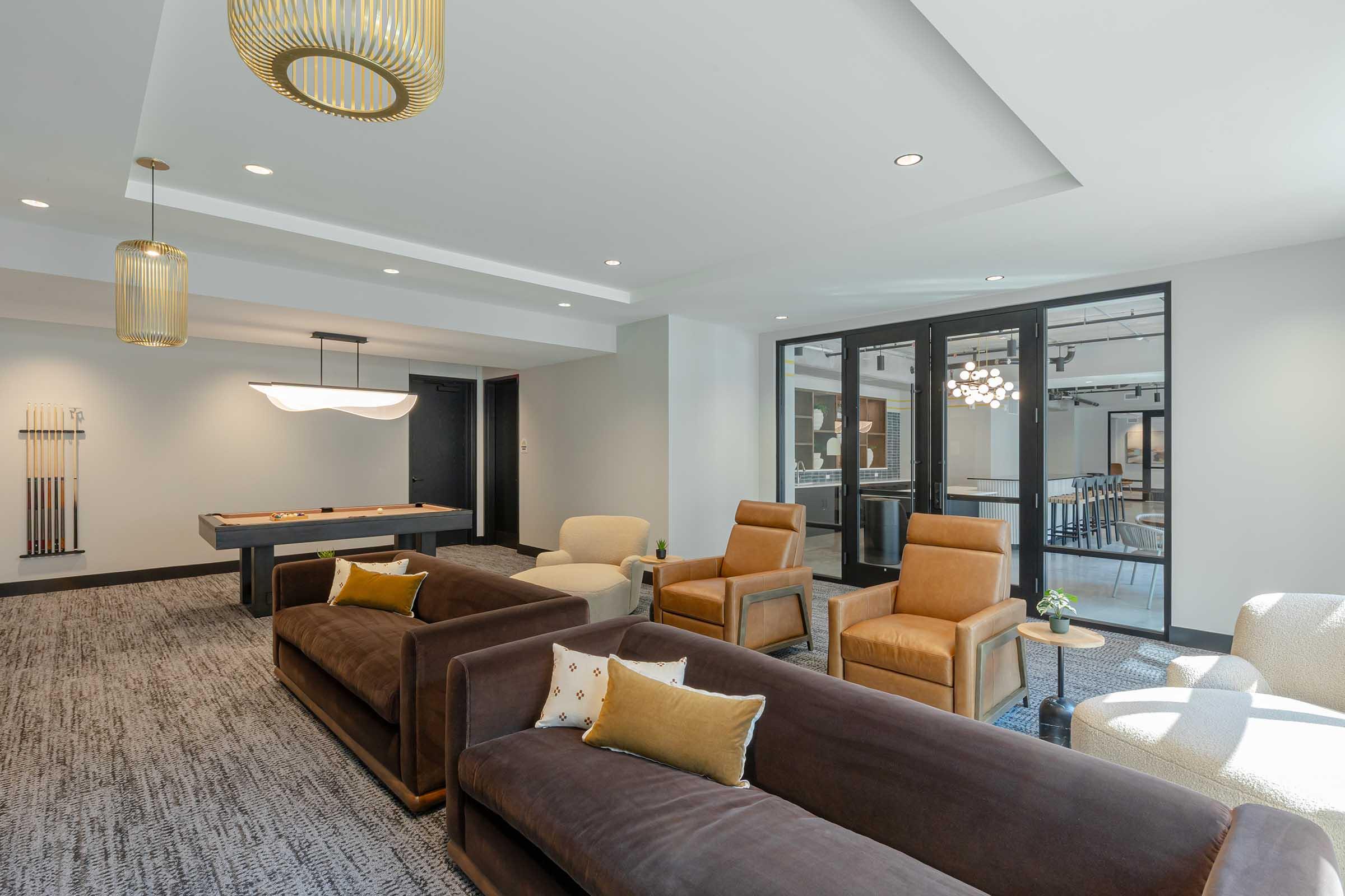 A modern lounge area featuring plush brown sofas and stylish armchairs. The space has a pool table in the background, large windows allowing natural light, and contemporary light fixtures. Decorative plants and cushions add a cozy touch to the inviting environment.