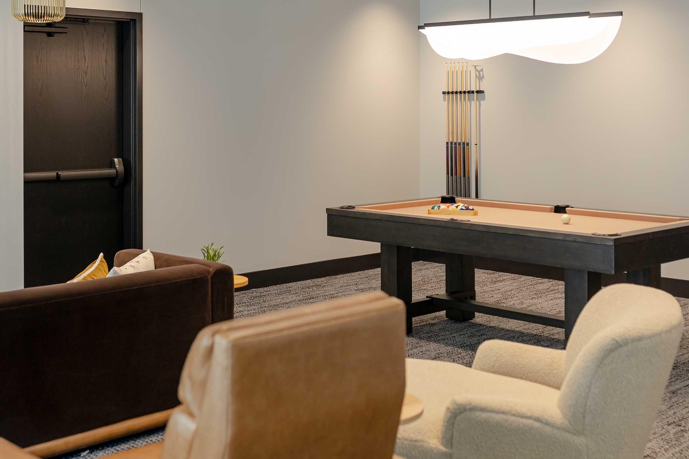 A cozy lounge area featuring a billiards table, two comfortable chairs, and a stylish light fixture. The walls are painted in neutral tones, and there’s a door leading to another room. A decorative element, such as a small plant or cushion, adds a warm touch to the space.