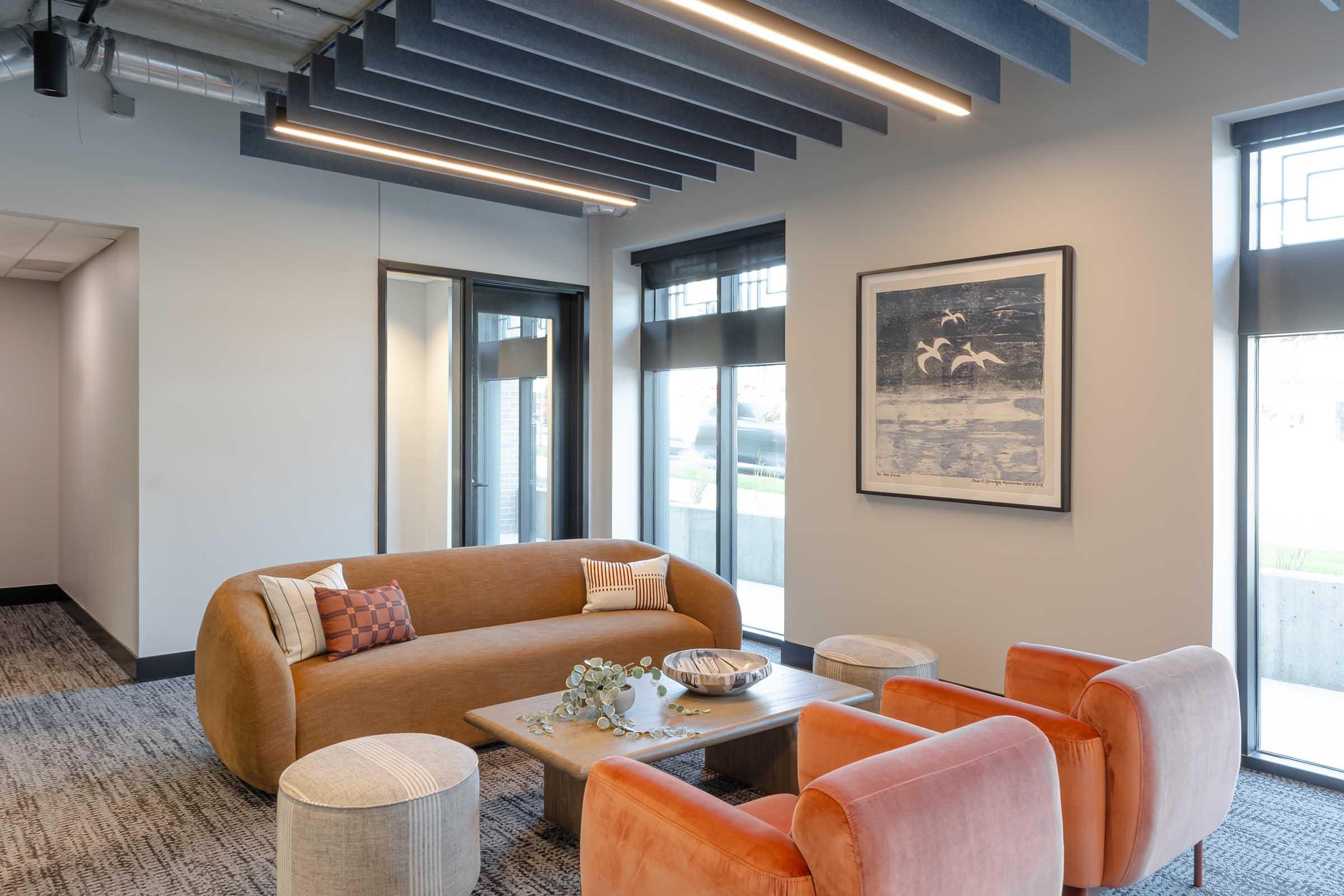 A modern lounge area featuring a curved orange sofa, two round orange chairs, and a wooden coffee table. The space is bright with natural light from large windows, and decorated with artwork and plants, creating a warm and inviting atmosphere.