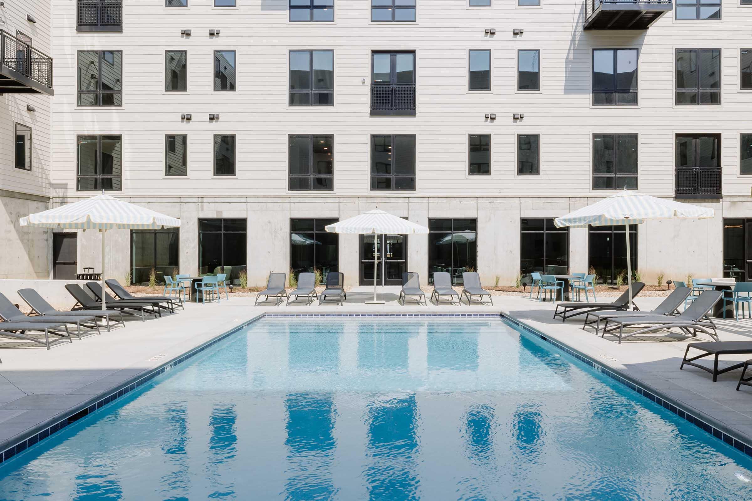 A modern outdoor swimming pool surrounded by lounge chairs and umbrellas. The pool reflects the building's facade, which features large windows and a contemporary design. The area is well-lit and inviting, perfect for relaxation and leisure.