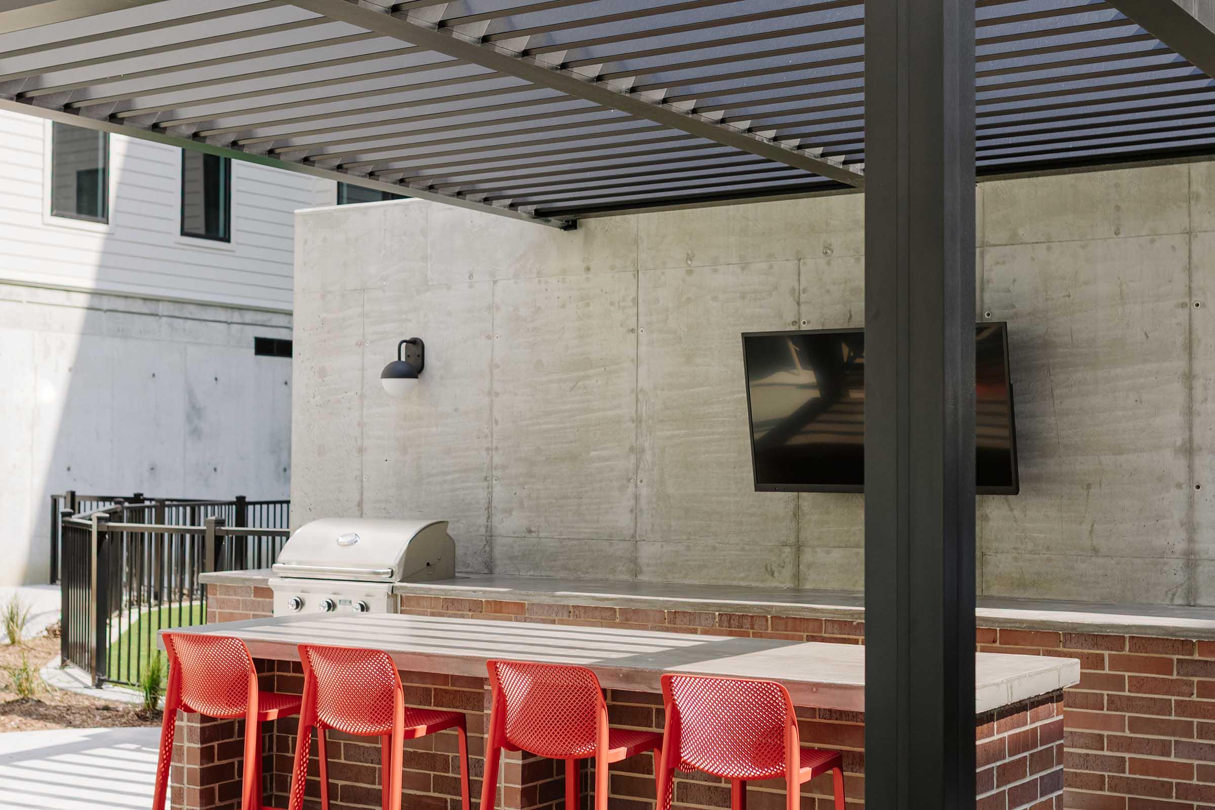 An outdoor patio area featuring a modern grill, a mounted television, and a long rectangular table with bright red chairs. The space is partially shaded by a metal pergola and surrounded by a concrete wall, providing a contemporary and inviting atmosphere for social gatherings.