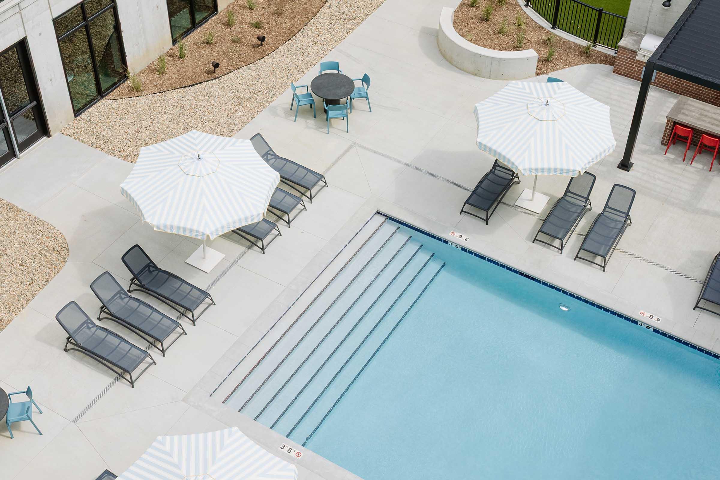 Aerial view of a modern swimming pool area featuring light blue water, grey lounge chairs, and large striped umbrellas. Surrounding the pool are landscaped areas with gravel and sparse plantings, along with a circular table and chairs, and a shaded pavilion nearby.