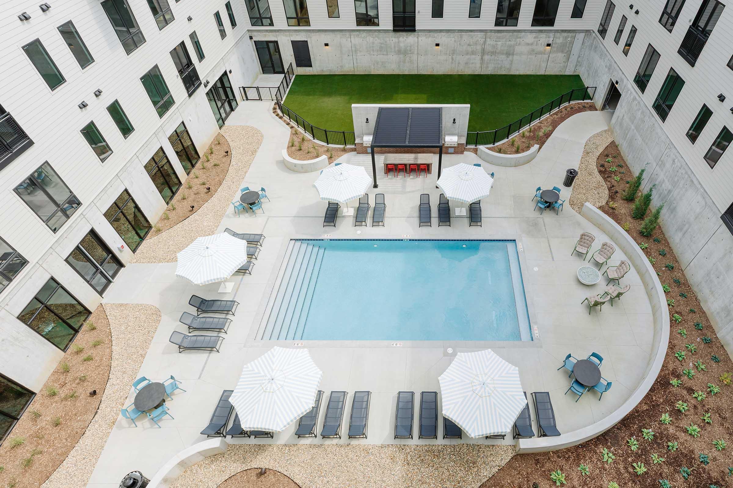 Aerial view of a modern apartment complex courtyard featuring a swimming pool surrounded by lounge chairs and umbrellas. Manicured landscaping includes gravel and green grass areas, with additional seating around the pool. The building has large windows and a contemporary design.