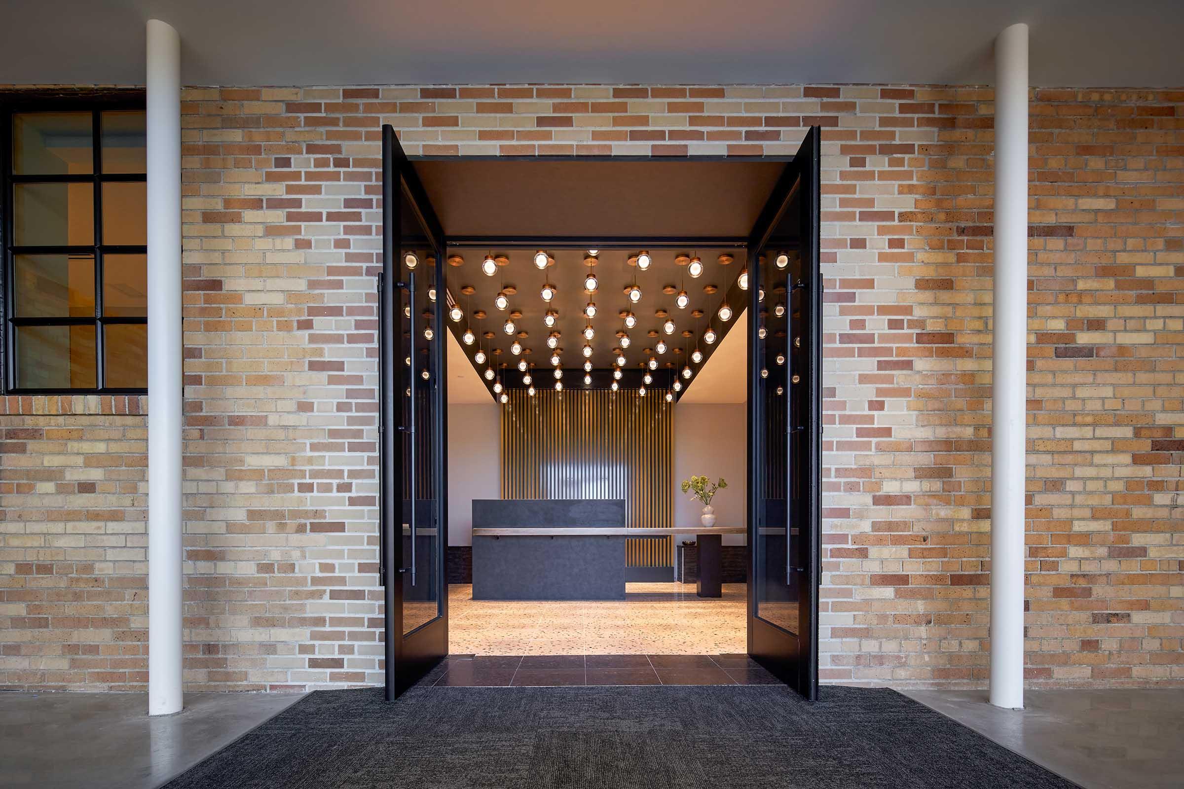 Wide open double doors leading into a modern interior space with a minimalist design. The entrance features a textured brick exterior and an illuminated ceiling with numerous light fixtures. Inside, there is a sleek reception desk and a simple decorative plant enhancing the contemporary atmosphere.