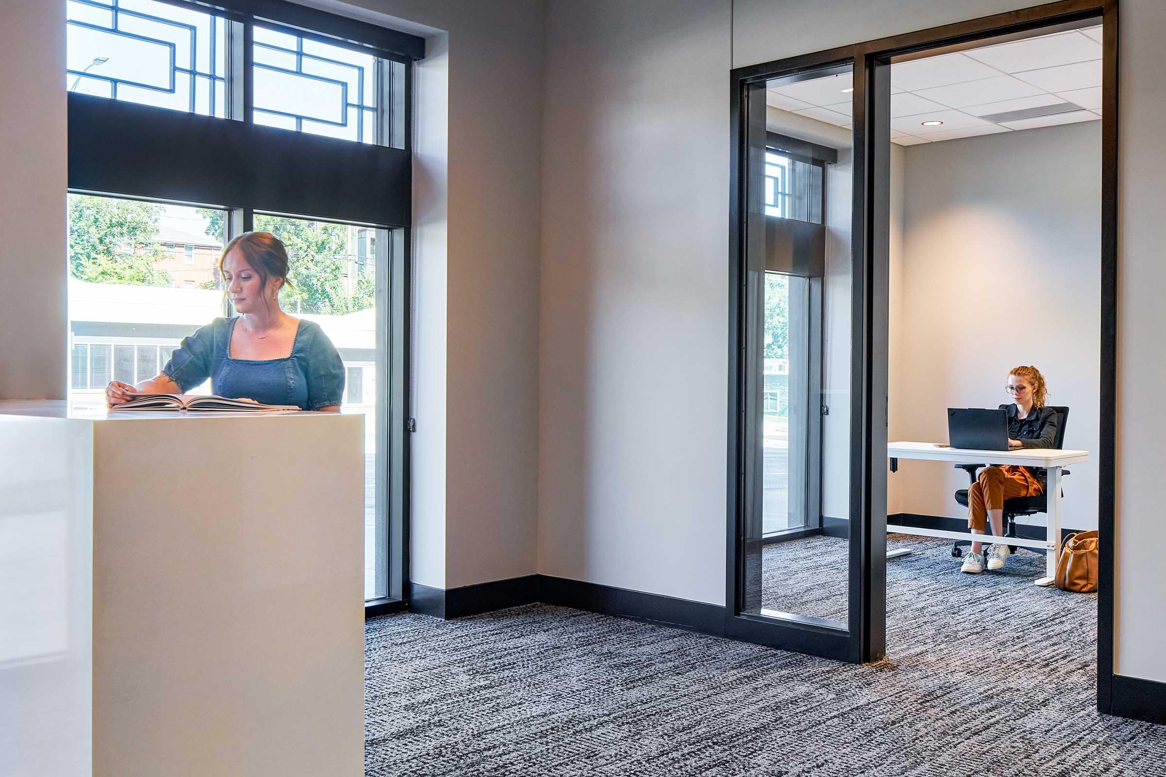 A modern indoor space featuring two women. One is reading a book near a large window, while the other sits at a desk in a glass-walled office, working on a laptop. The room has neutral-colored walls and carpeting, with good natural light.