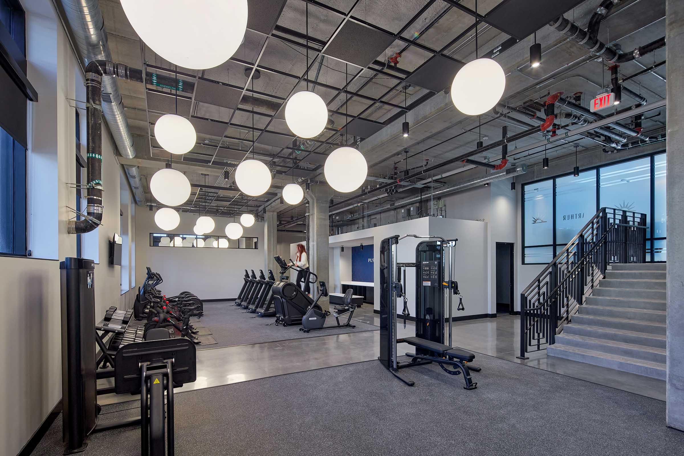A modern gym interior featuring fitness equipment like elliptical machines and weights. The space is well-lit with large round pendant lights and has an open layout, including a staircase leading to a second level. The floors are finished with a smooth surface and walls have a clean design.