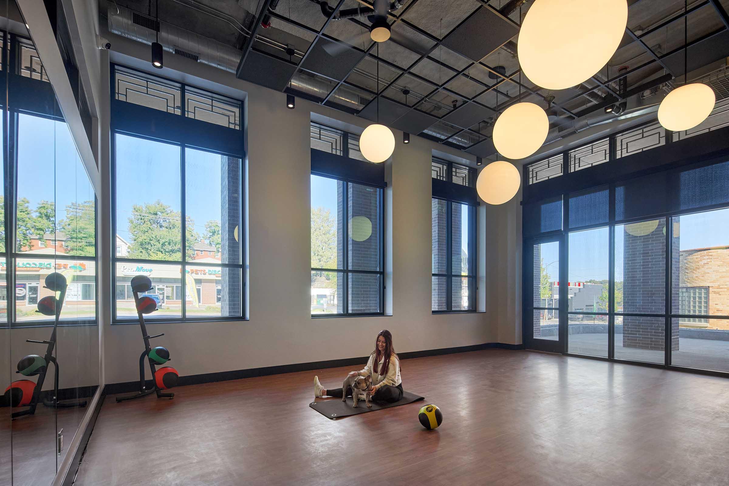 A spacious empty exercise room with large windows allowing natural light, featuring round pendant lights on the ceiling. A woman sits on a yoga mat with a dog beside her, and exercise equipment is visible in the corner. The view outside shows a street with shops.