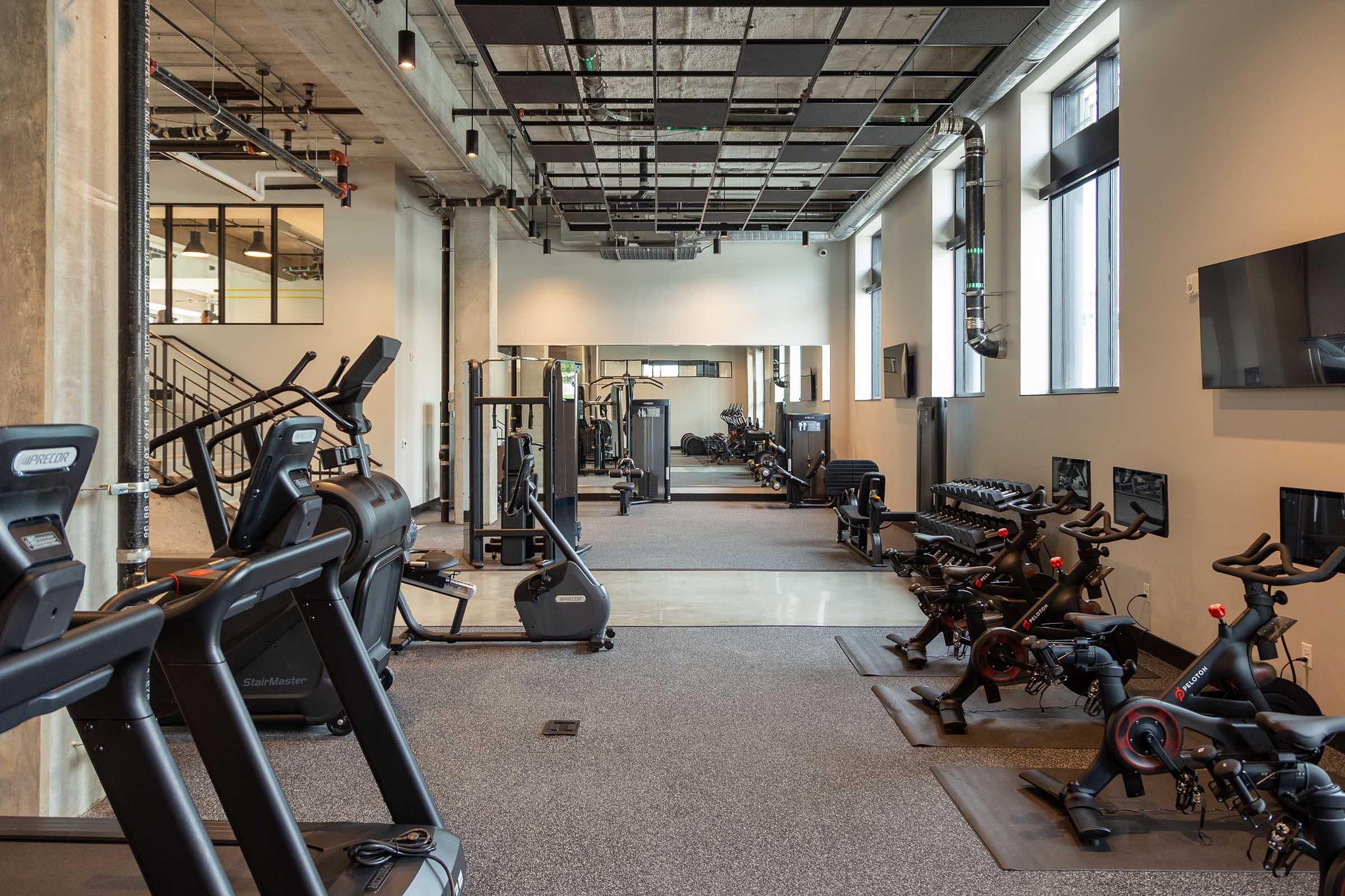 A modern gym interior featuring various exercise equipment such as treadmills, stationary bikes, weight benches, and free weights. The space has large windows for natural light, exposed ceiling beams, and a clean, minimalist design.