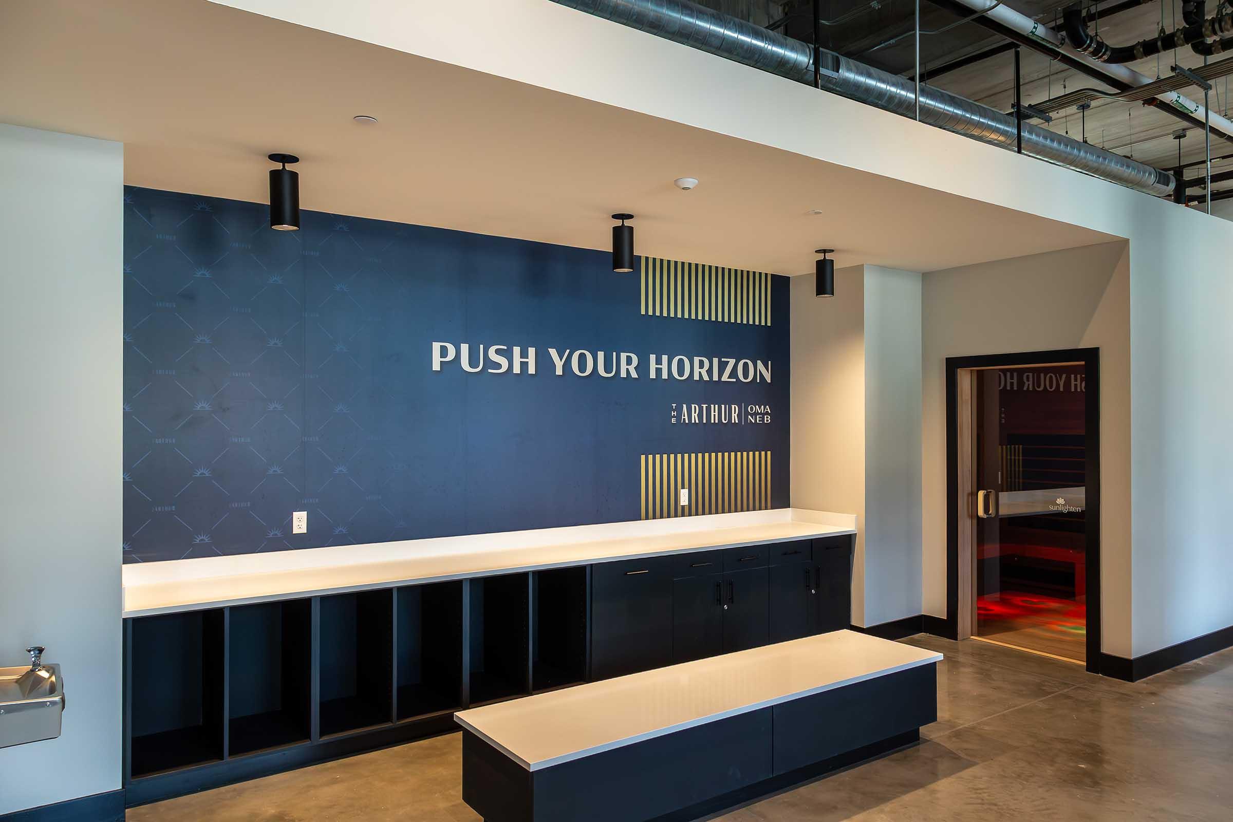 Interior of a modern lobby featuring a blue accent wall with the text "PUSH YOUR HORIZON" prominently displayed. There is a white countertop with seating and a water fountain. The space has a sleek, contemporary design with industrial elements and a doorway visible in the background.