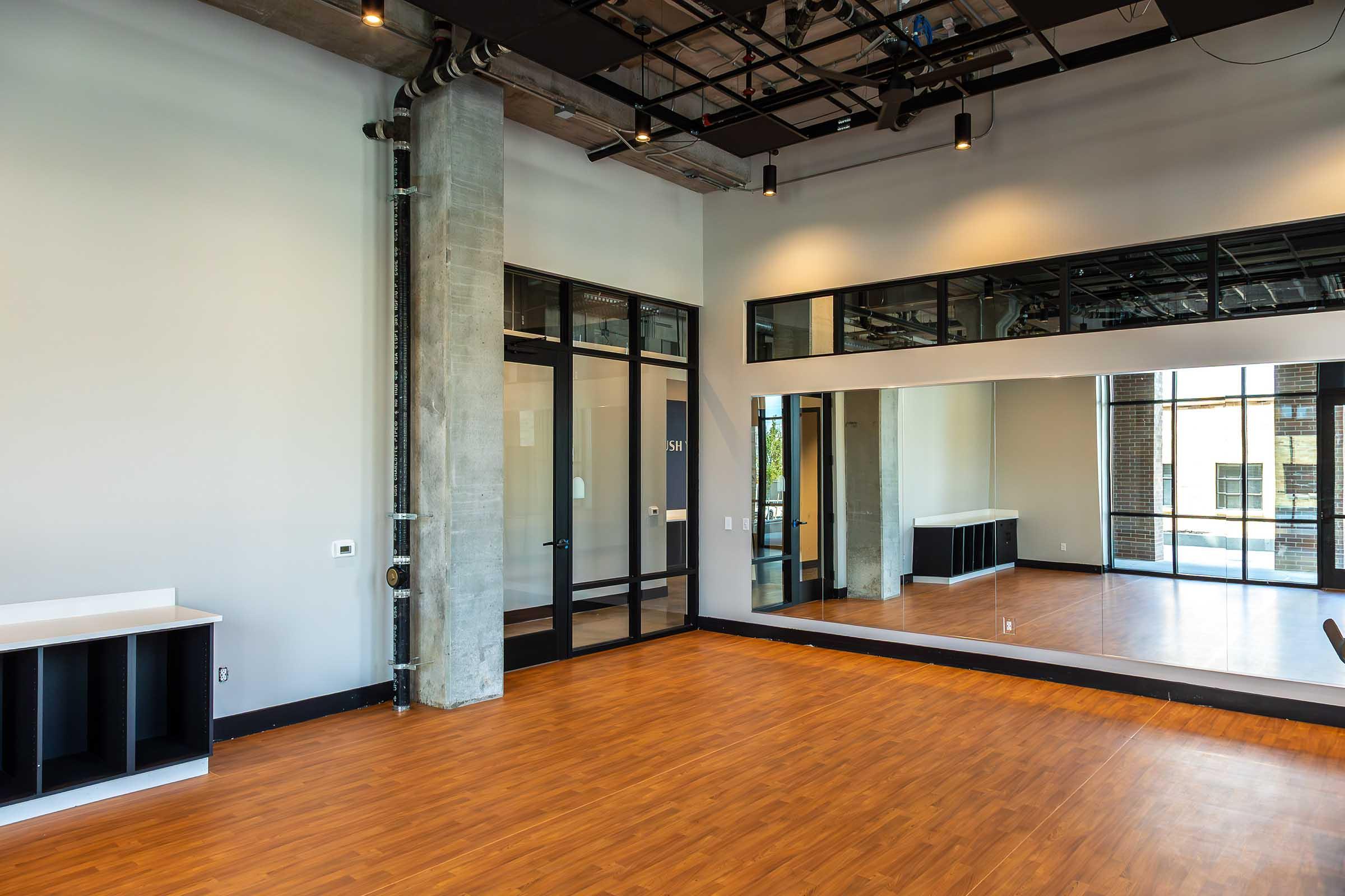 Spacious, modern interior of a studio or dance space featuring large windows, a mirrored wall, hardwood floors, and minimalist decor. The walls are light-colored, and there are exposed ductwork and industrial-style lighting fixtures.