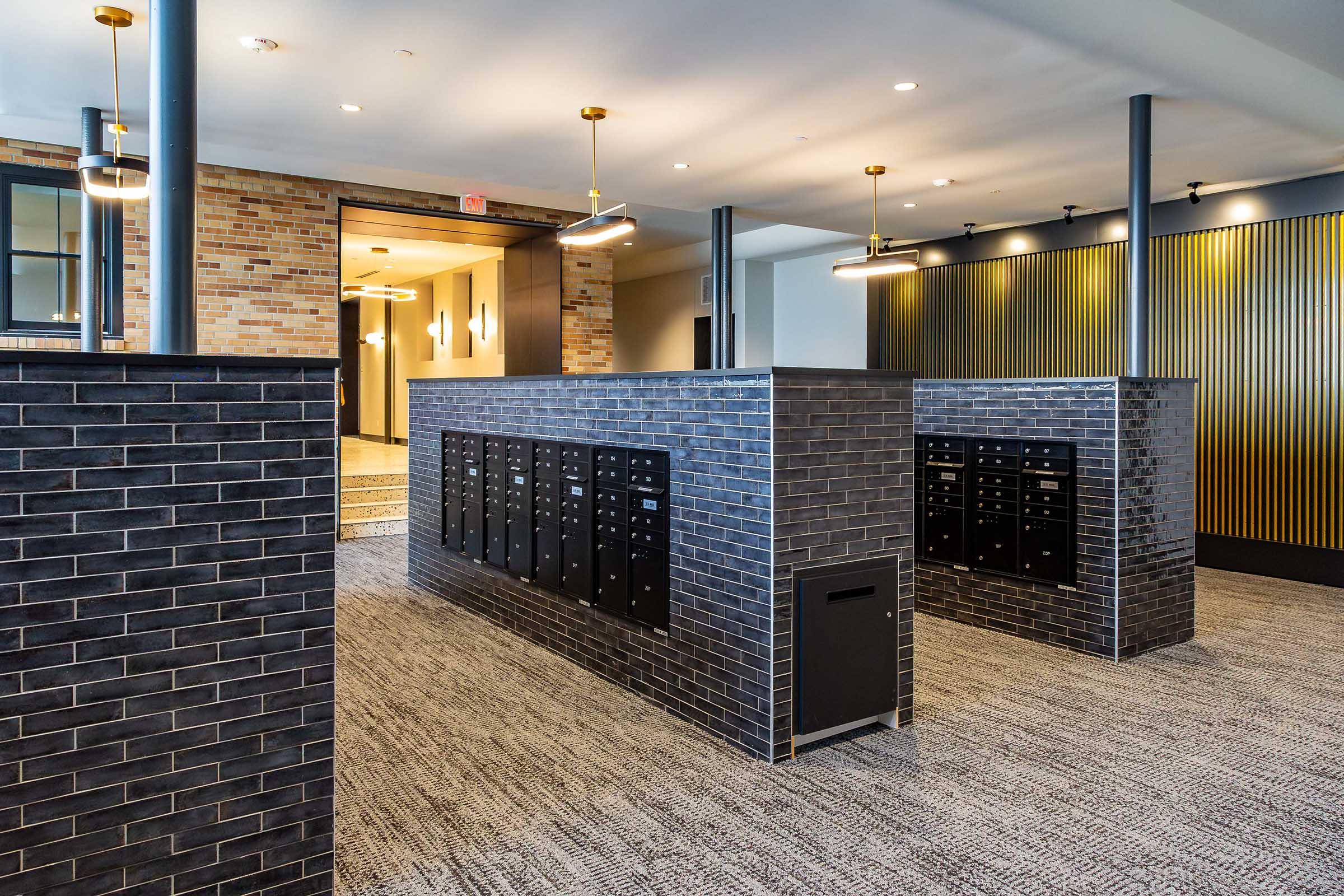 A modern lobby area featuring black brick walls and various mailboxes. The space is well-lit with pendant lights, and has a stylish carpeted floor. There's a decorative partition creating sections, with a sleek, contemporary design that blends functionality and aesthetics.