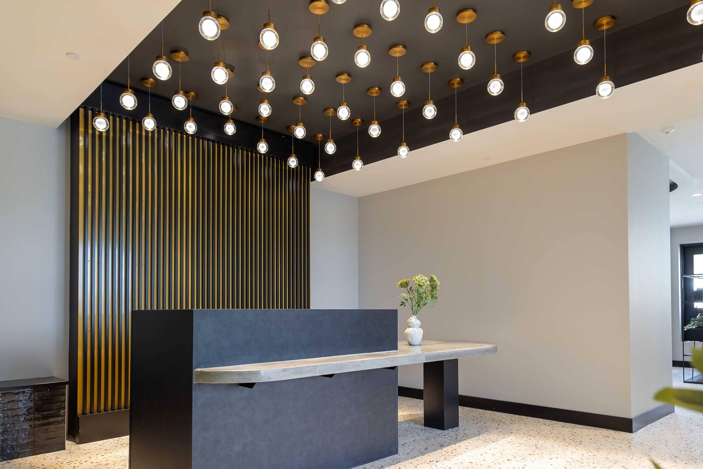 A modern lobby featuring a sleek reception desk made of dark material, a decorative wall with vertical gold stripes, and a unique ceiling adorned with multiple pendant lights hanging in an artistic arrangement. A vase with flowers adds a touch of nature to the contemporary space.