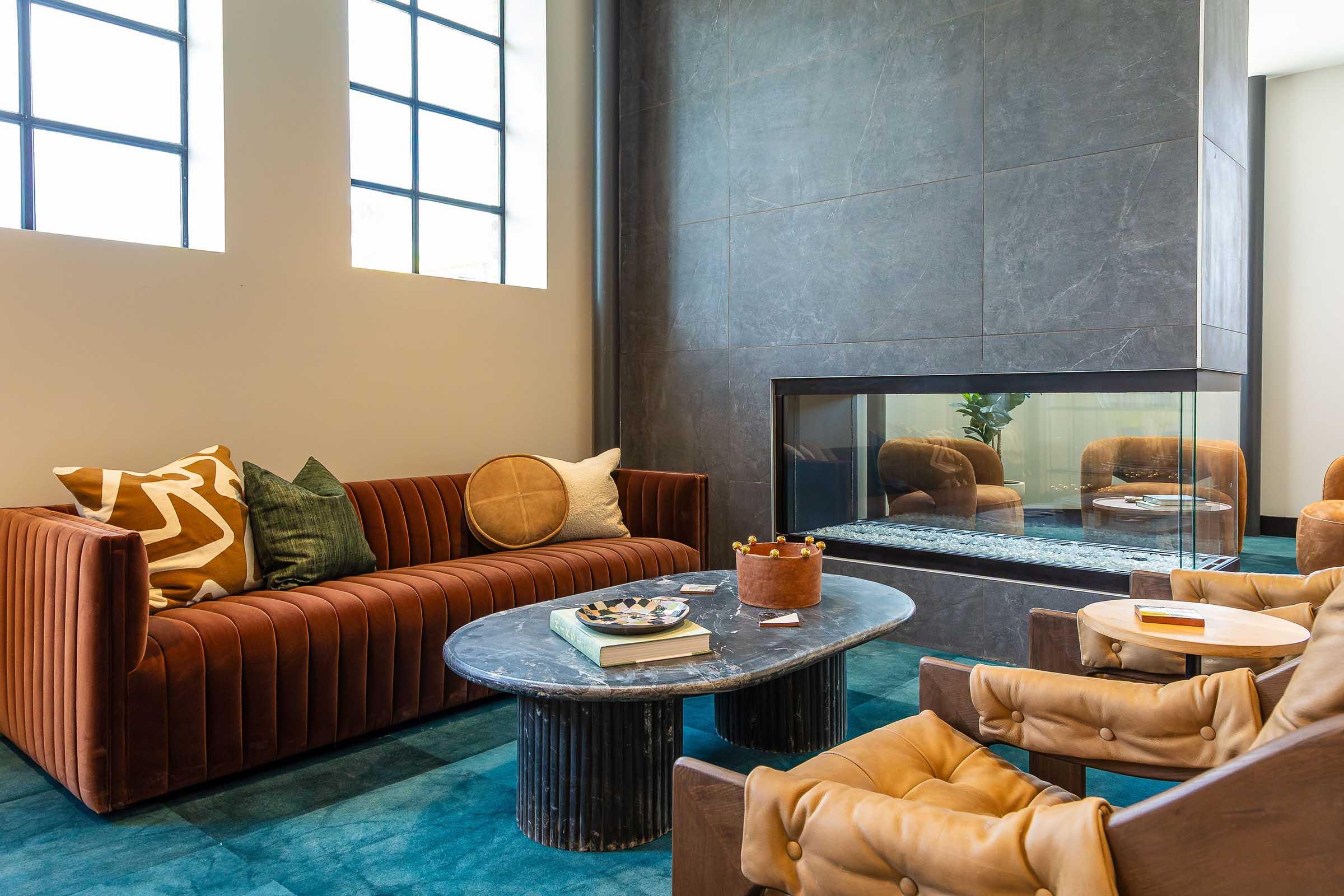 A modern living room featuring a cozy brown sofa adorned with decorative pillows, a round decorative pillow, and an elegant glass fireplace. The space includes a circular coffee table with decorative items, and plush brown armchairs. Natural light streams in through large windows, enhancing the inviting atmosphere.