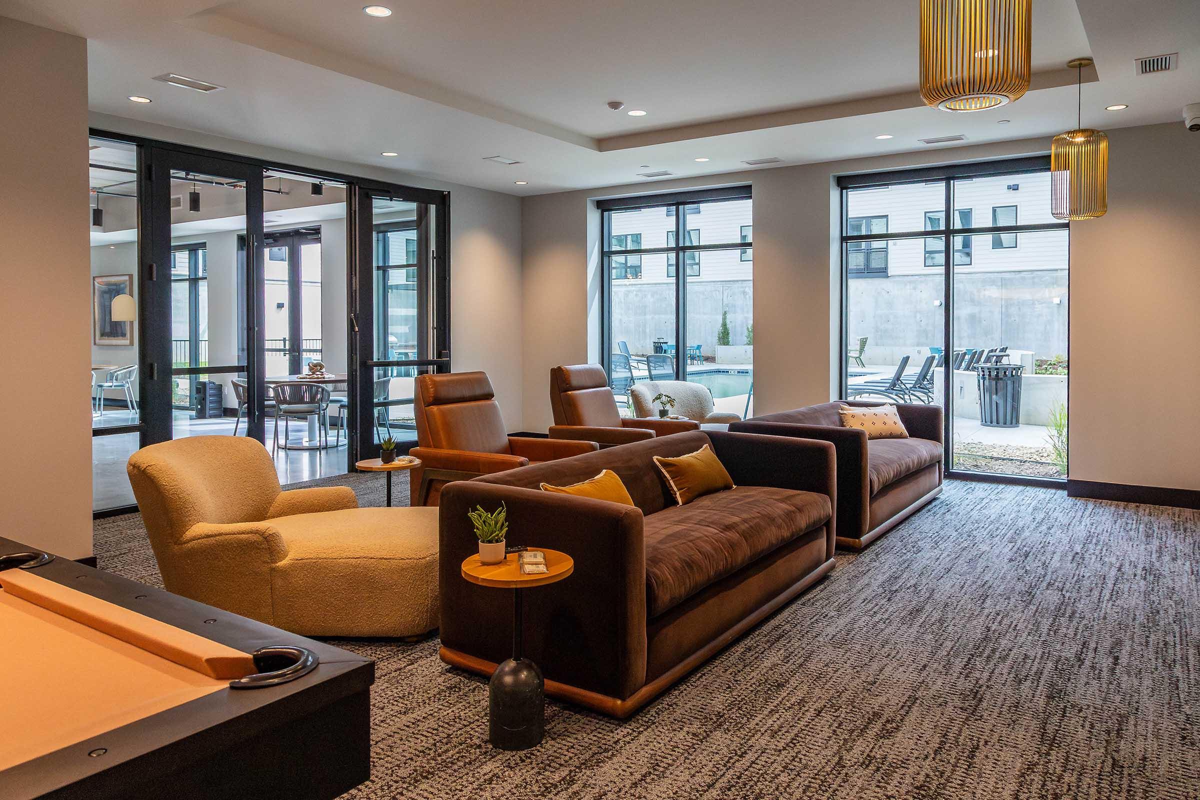 A modern lounge area featuring two plush sofas and an armchair, with large windows allowing natural light, a small plant on the coffee table, and a view of an outdoor seating area. The decor is stylish and contemporary, designed for relaxation and socializing. A pool table is visible in the foreground.