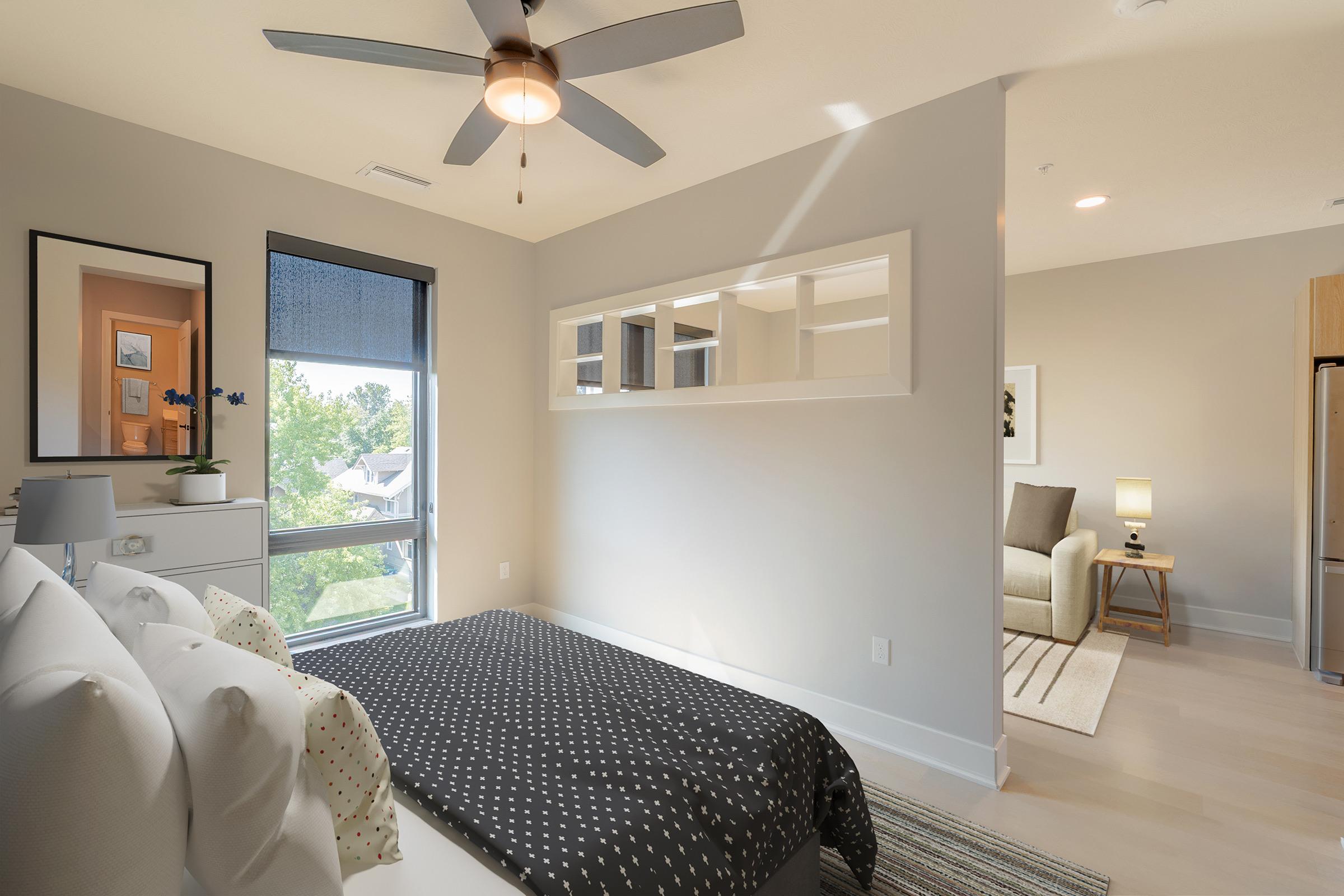 A modern bedroom featuring a queen-sized bed with a polka dot comforter, a ceiling fan, and a large window allowing natural light. A mirror and a small decorative plant are on the nightstand, and a cozy seating area is visible in the background with a chair and lamp. The walls are painted in neutral tones.