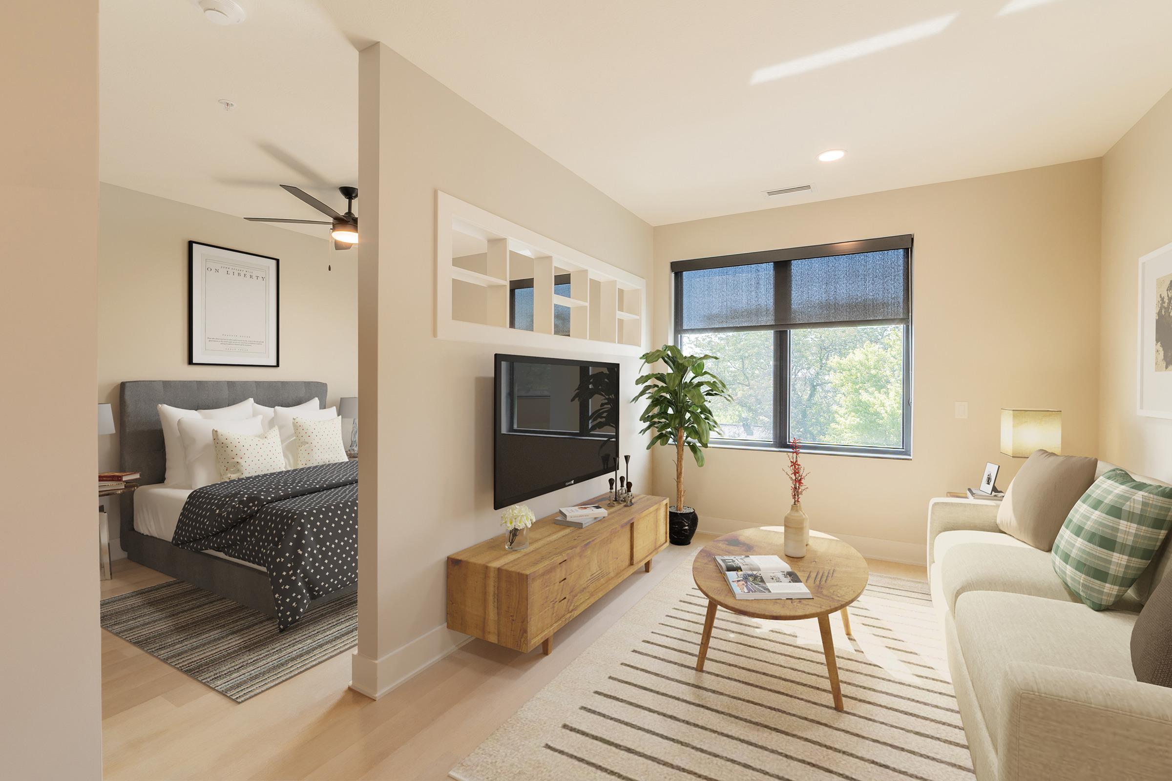 Bright and modern living space featuring a cozy seating area with a light-colored sofa, circular coffee table, and a decorative plant. A flat-screen TV is mounted on the wall above a wooden TV stand. The adjoining bedroom is visible, with a stylish bed and artwork enhancing the inviting atmosphere.