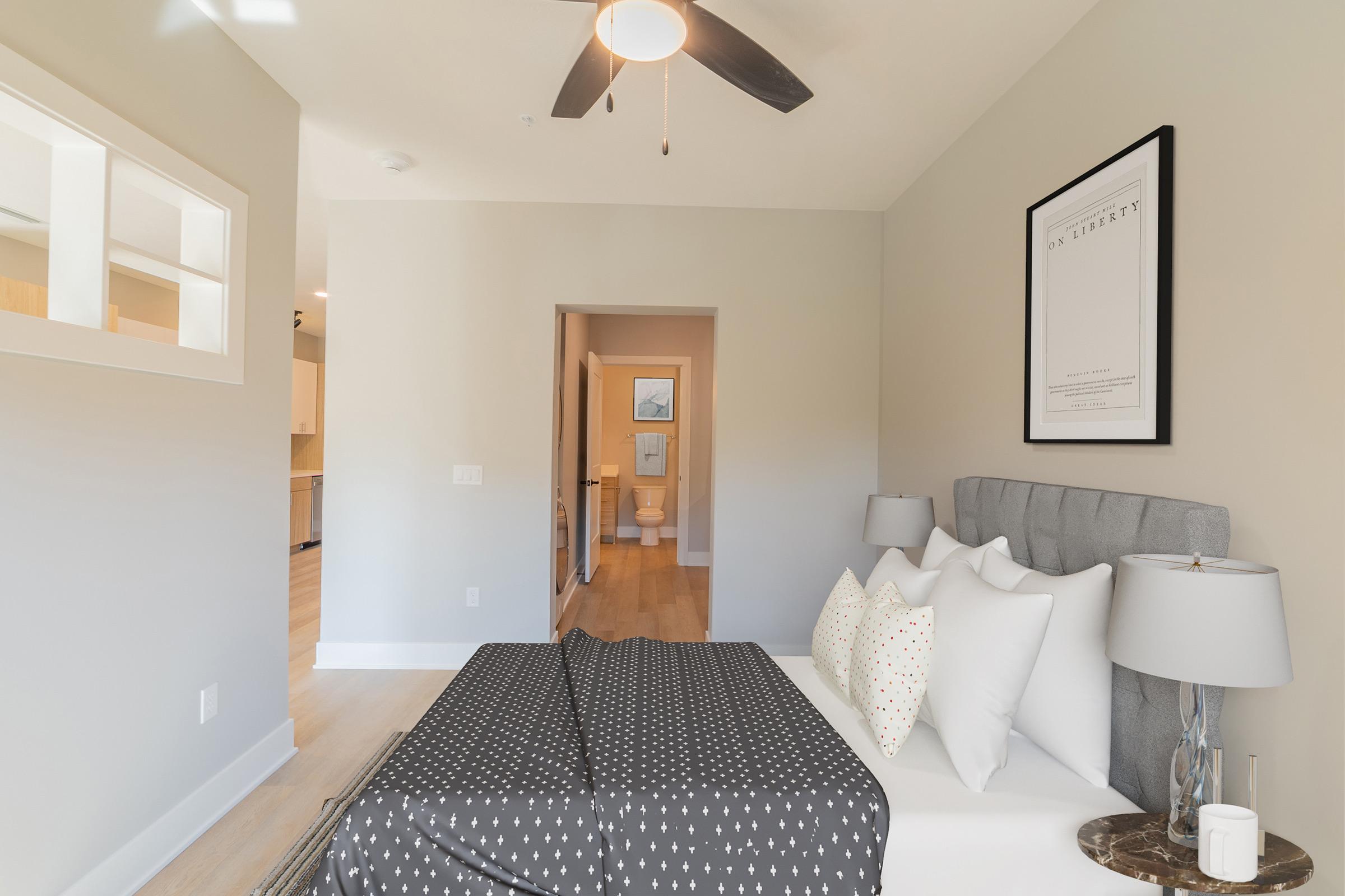 A modern bedroom featuring a gray upholstered bed with white pillows and a patterned blanket. A decorative lamp sits on a nightstand. In the background, a doorway leads to a well-lit hallway and a bathroom, showcasing light wood flooring and neutral-colored walls.