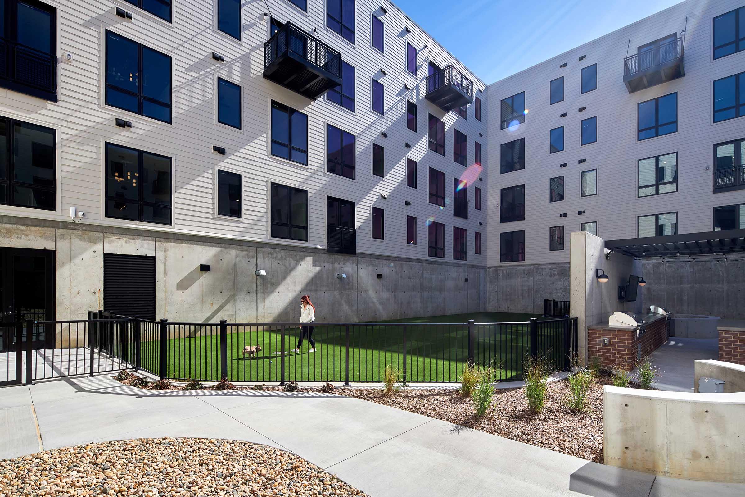A modern apartment complex courtyard featuring well-maintained grass, a fenced area where a person walks a small dog, and contemporary architecture with large windows and balconies. The space is bright with sunlight, showcasing a blend of concrete and landscaping elements.