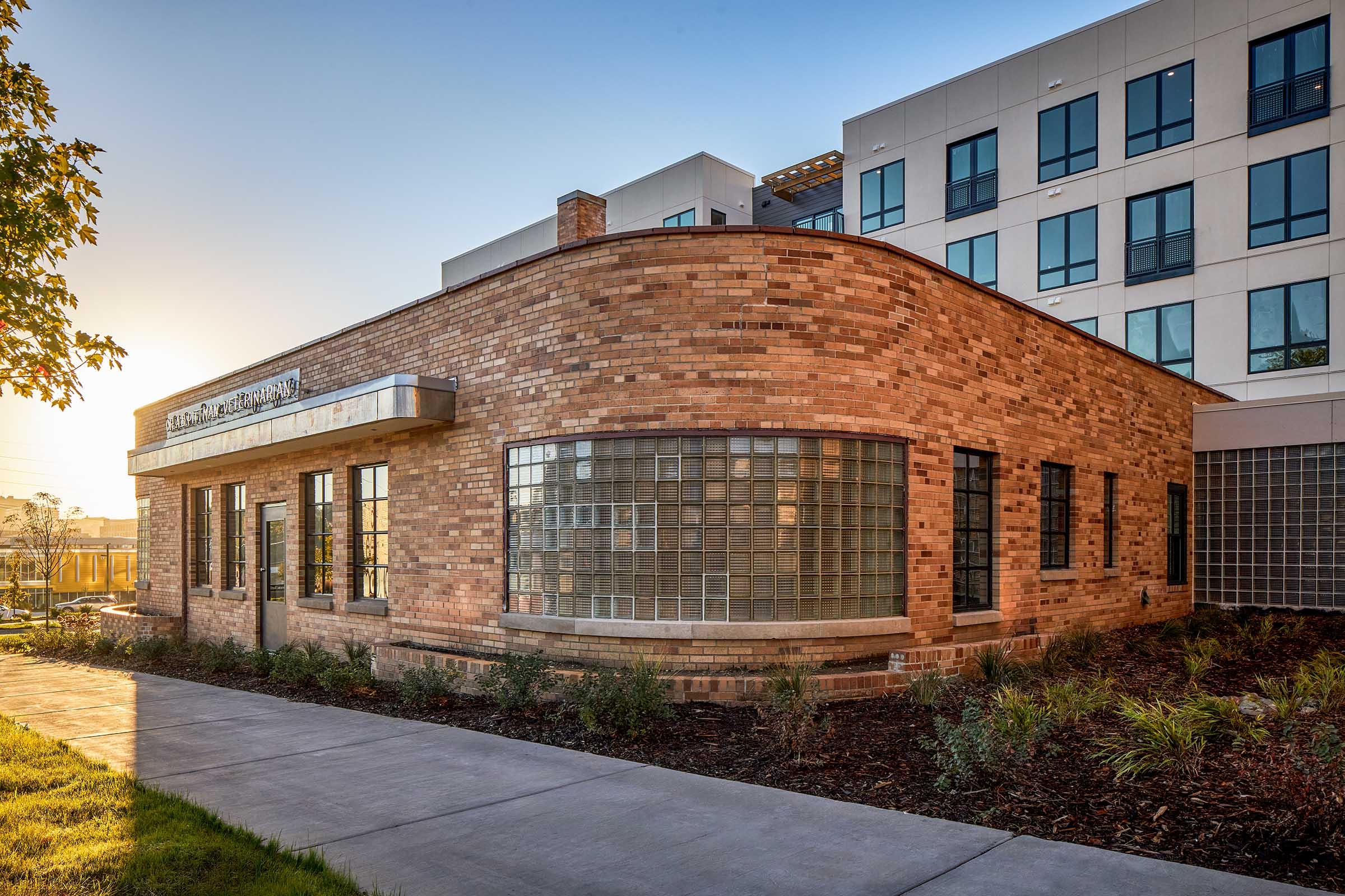 A modern brick building with large glass block windows, featuring an arched entrance. The facade is complemented by a landscaped area with greenery. In the background, a contemporary multi-story building is visible, bathed in warm light from a sunset. The scene exudes a blend of historic and modern architecture.