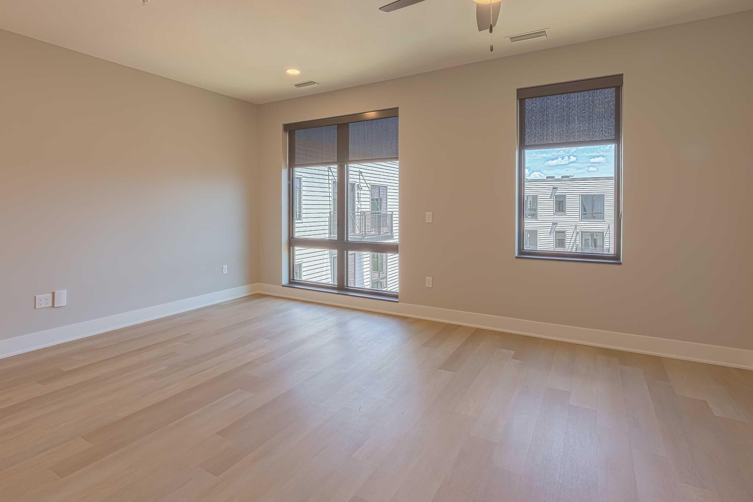 A spacious empty room with light-colored wooden flooring, two large windows with dark blinds, and a ceiling fan. The walls are painted in a neutral tone, creating a bright and airy atmosphere. Suitable for various interior design possibilities.