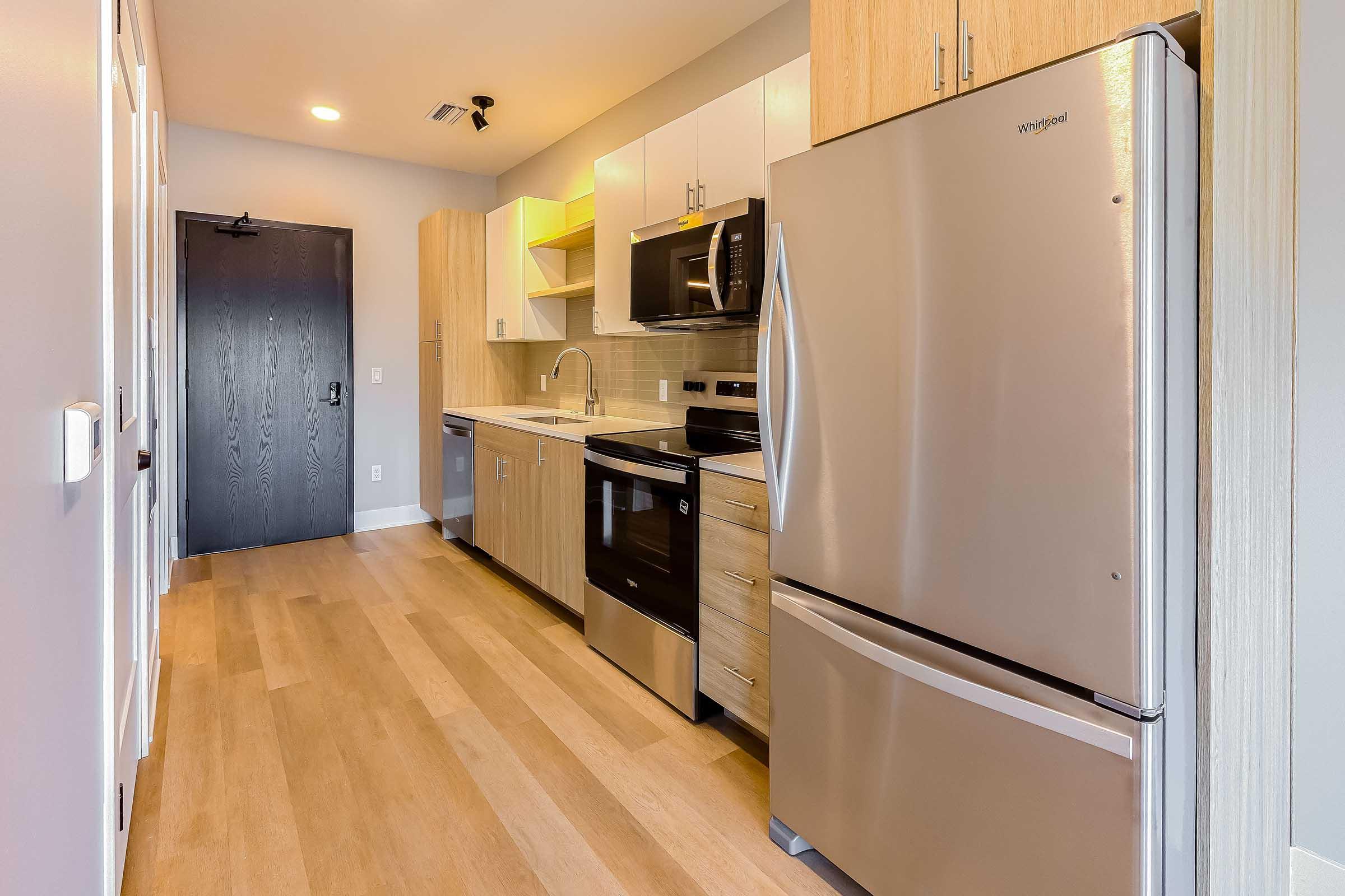 Modern kitchen featuring stainless steel appliances, including a refrigerator, microwave, and dishwasher. Light wood cabinets and countertops provide a warm aesthetic. The space has a bright and open layout with natural light illuminating the area, and a door to the left leading outside.