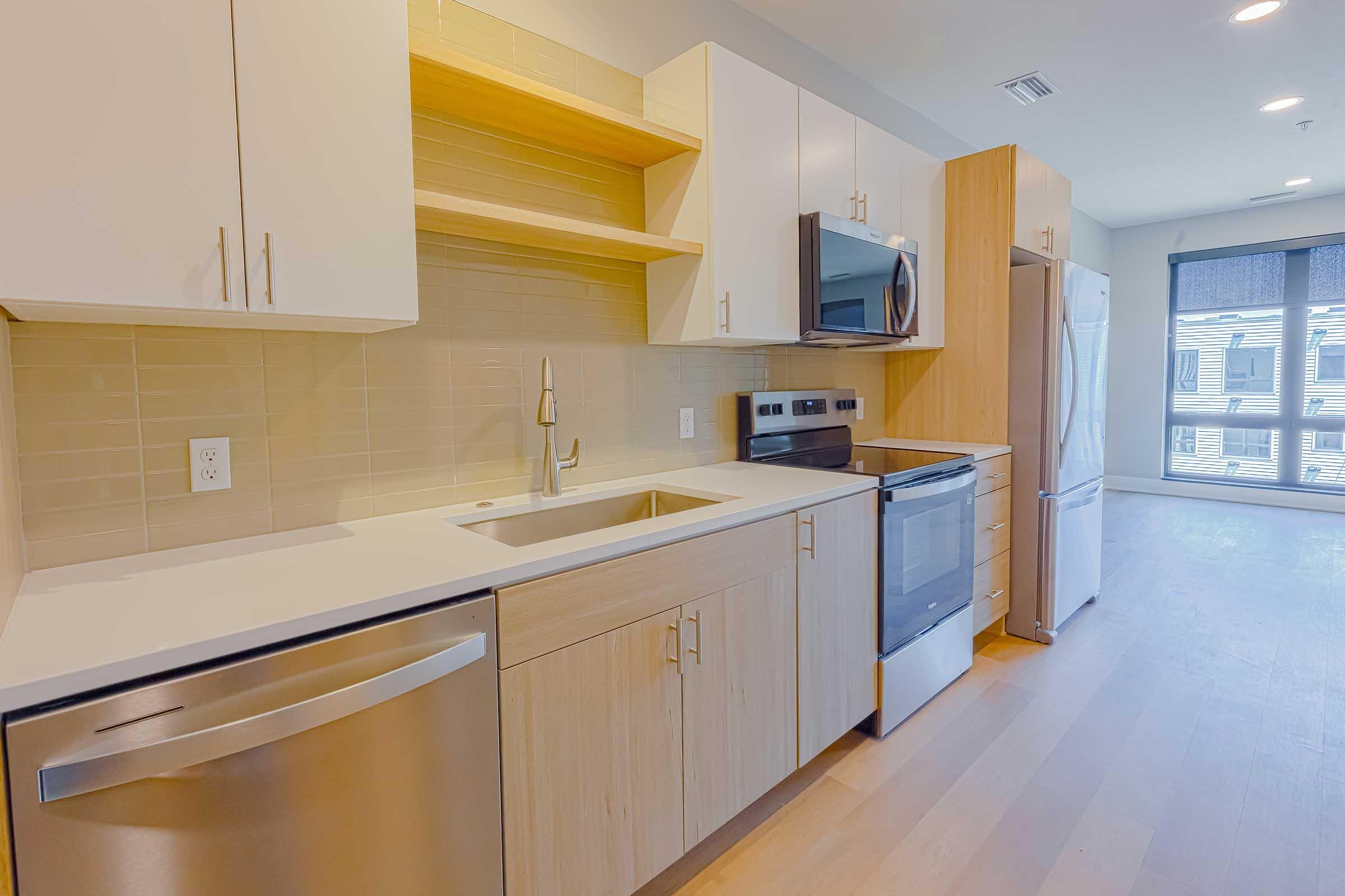 Modern kitchen featuring white and light wood cabinets, stainless steel appliances including a microwave, oven, and refrigerator. The sink is integrated into the countertop, with a pull-down faucet. Natural light streams in through a window with blinds, creating a bright and inviting atmosphere.