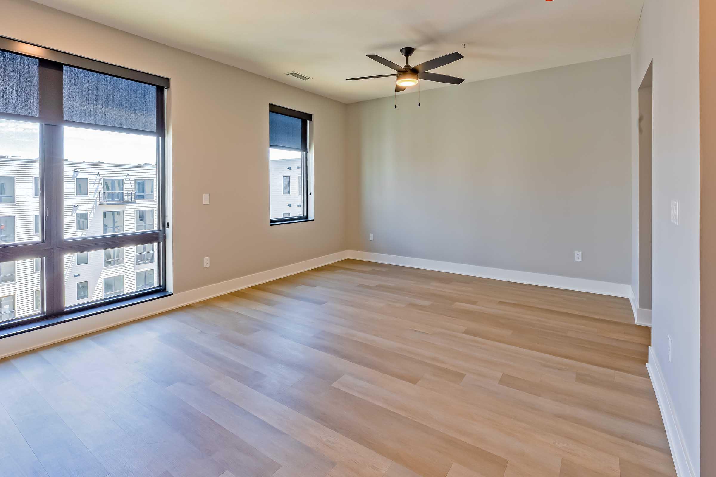 A spacious, empty room with light-colored wooden flooring, large windows featuring black roller shades, and a ceiling fan. The walls are painted in a neutral tone, creating a bright and open environment. An adjoining doorway leads to another area, enhancing the room's layout.