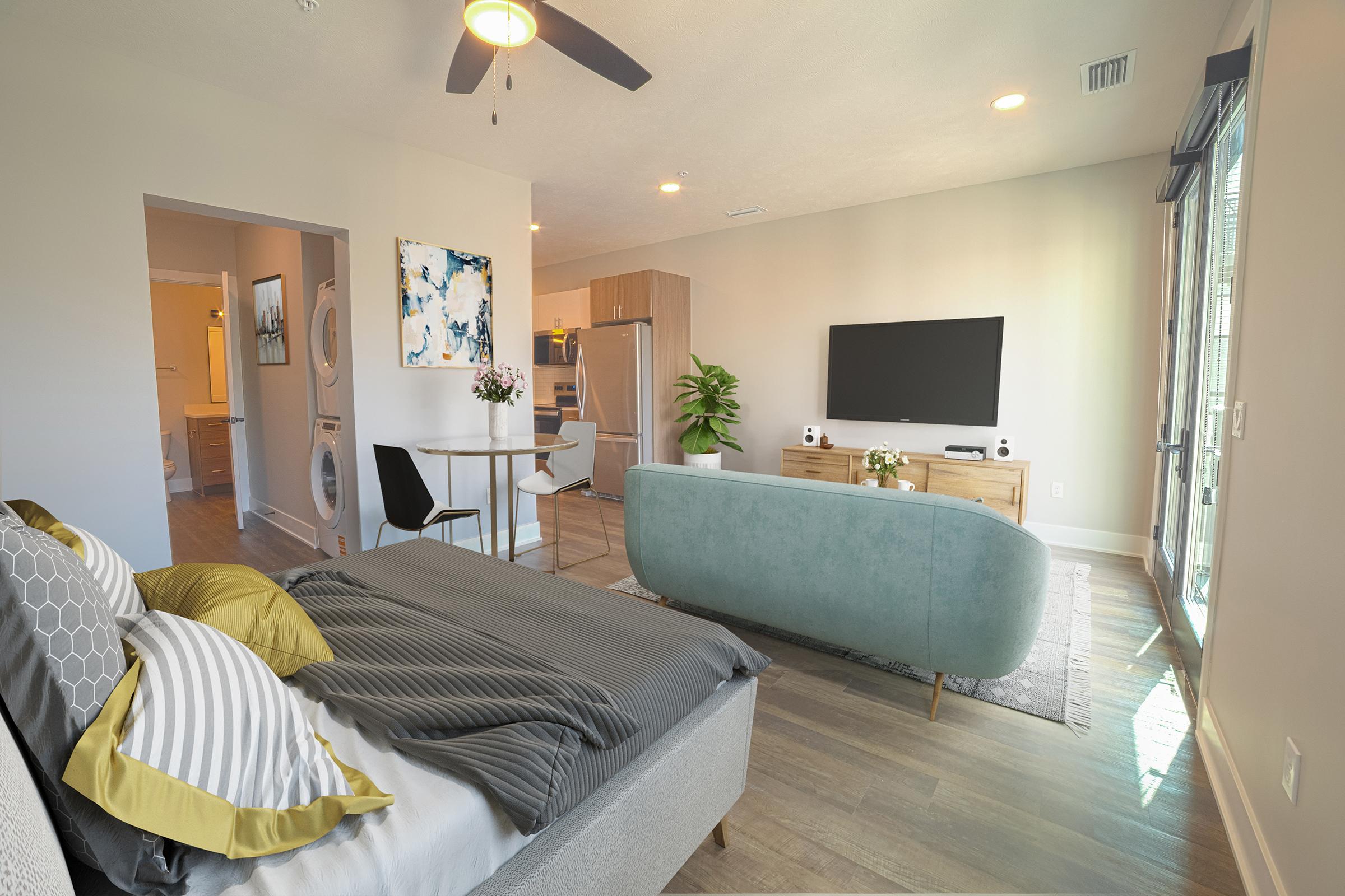 A modern studio apartment featuring a cozy bedroom with a bed adorned in gray and yellow pillows. The living area includes a green sofa, a wooden TV stand with a flat-screen TV, and a kitchen area visible in the background. Large windows provide natural light, and decorative plants add a touch of greenery.