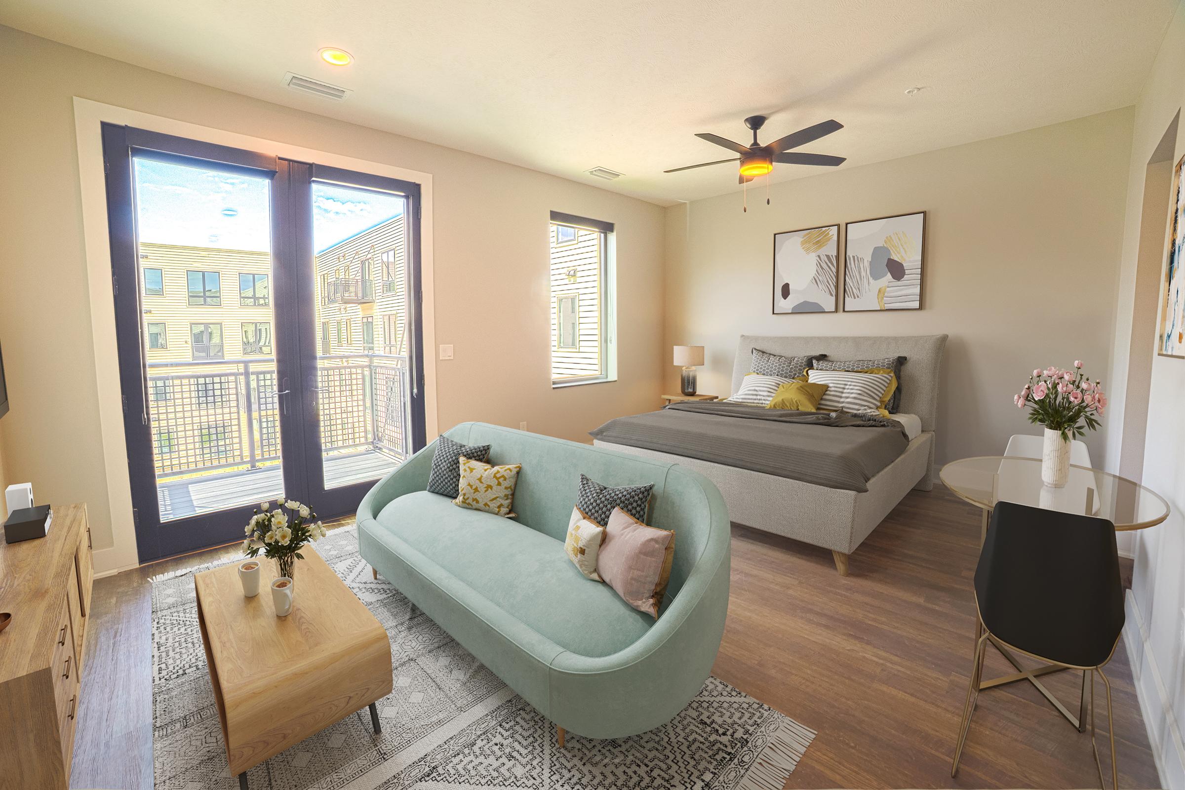 A modern, spacious bedroom and living area featuring a large bed with gray bedding, a light blue sofa, a wooden coffee table, and a stylish side table. The room has large windows revealing a view of an outdoor balcony, brightening the space with natural light. Decor includes wall art and a small vase of flowers.