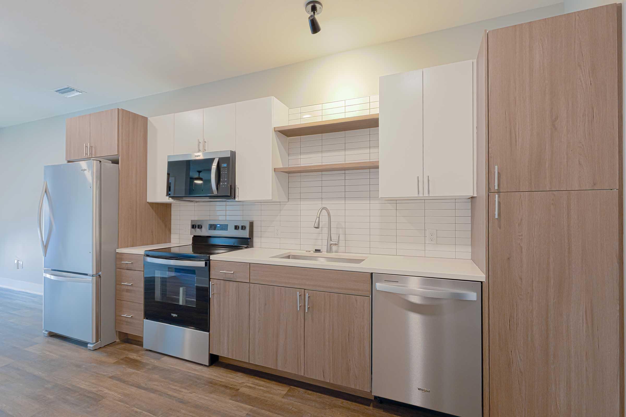 Modern kitchen featuring stainless steel appliances, including a refrigerator, microwave, stove, and dishwasher. The cabinetry combines wood and white finishes, with an open shelving area above the countertop. The backsplash consists of white tiles, and the flooring is a warm wood tone, creating a contemporary aesthetic.