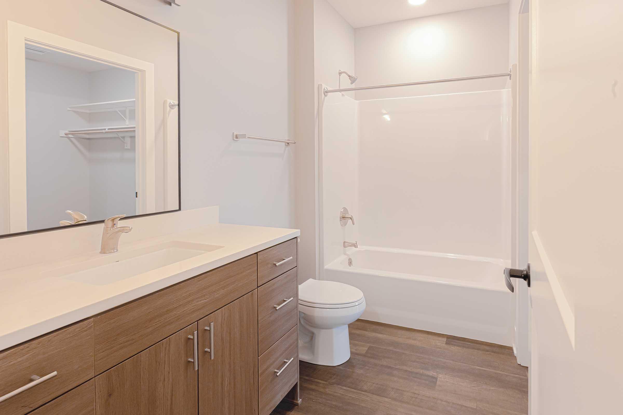 A modern bathroom featuring a white toilet next to a small vanity with a sink, set against light-colored walls. The space includes a shower with glass doors and shelving built into the wall. The flooring is a warm wood tone, enhancing the contemporary design.